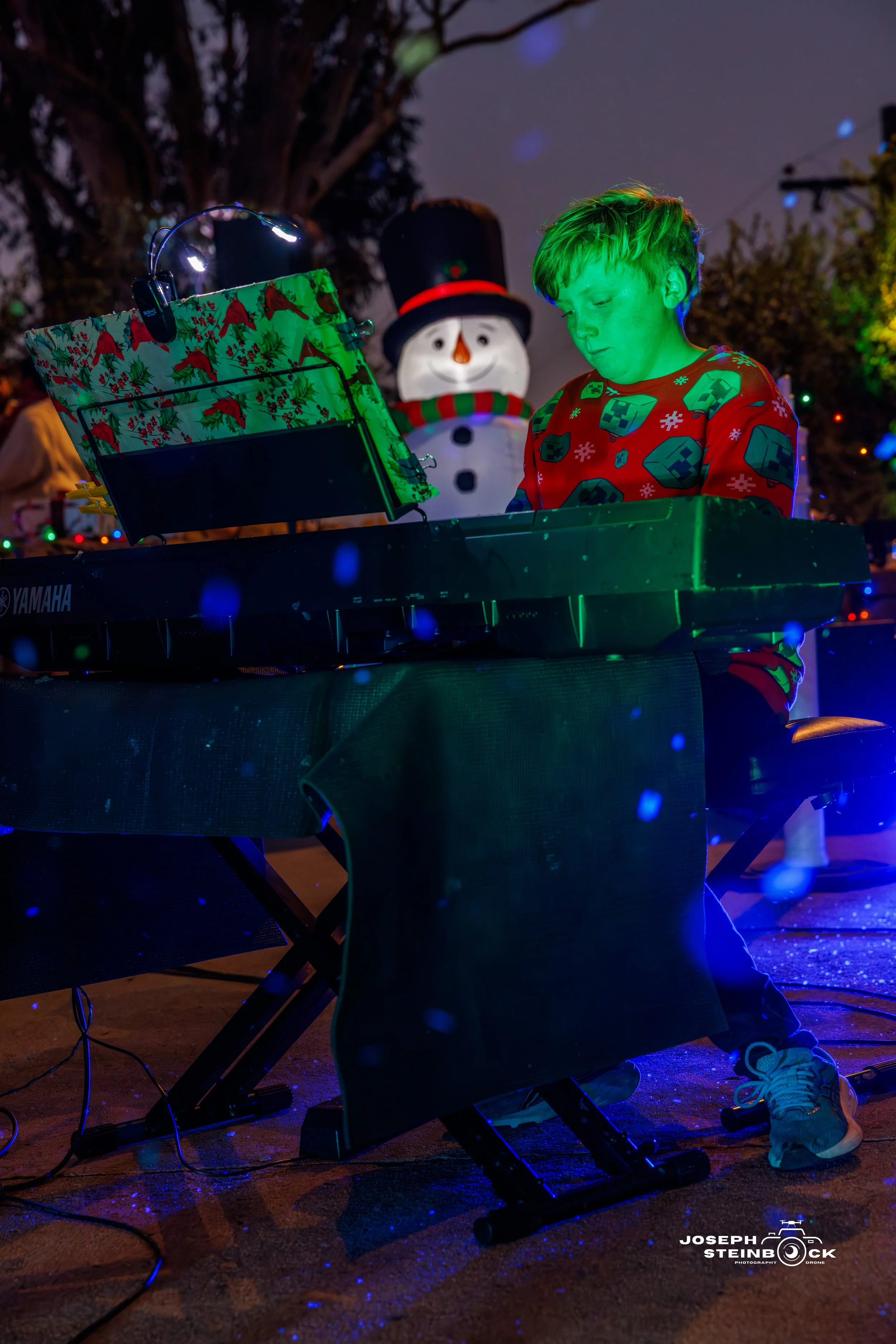 A young boy with green-tinted face playing a keyboard in a festive outdoor setting, decorated with Christmas lights, a snowman decoration, and a wrapped gift box on the keyboard.