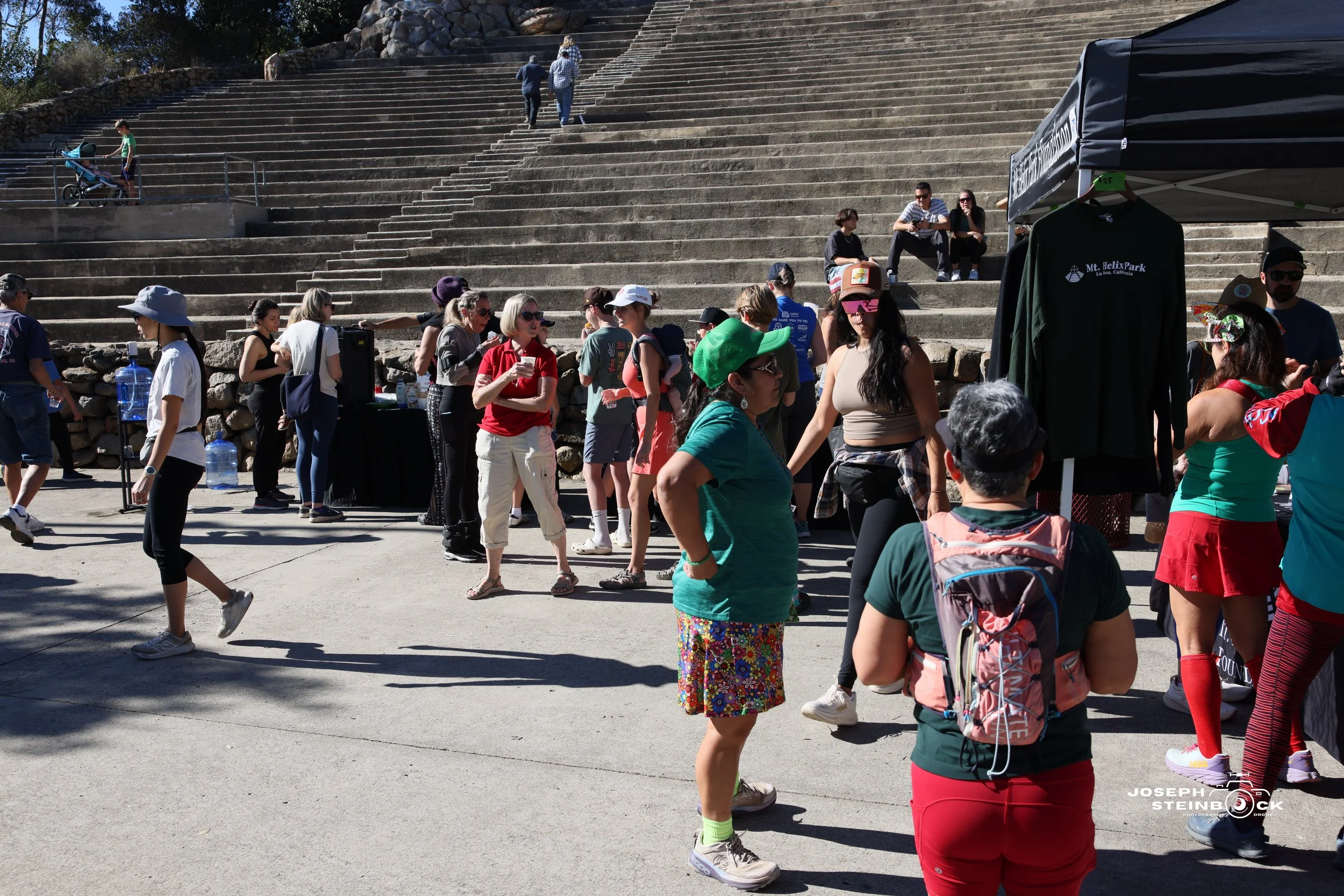 People gathering at an outdoor event near concrete stairs, with some browsing merchandise at a booth and others walking or sitting on the stairs.