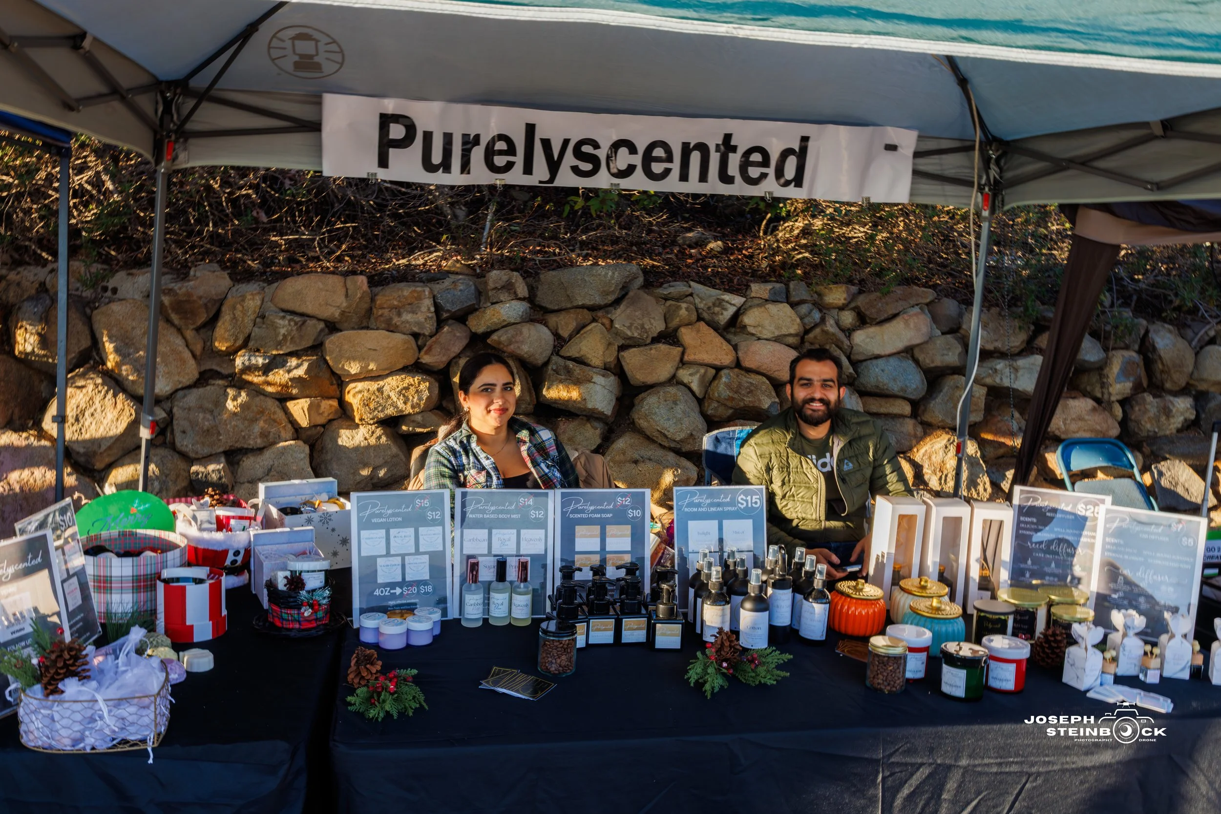Two people at a table selling scented products at an outdoor market stall with a 'Purely scented' banner, various candles, sprays, and gift items displayed in front of a stone wall.