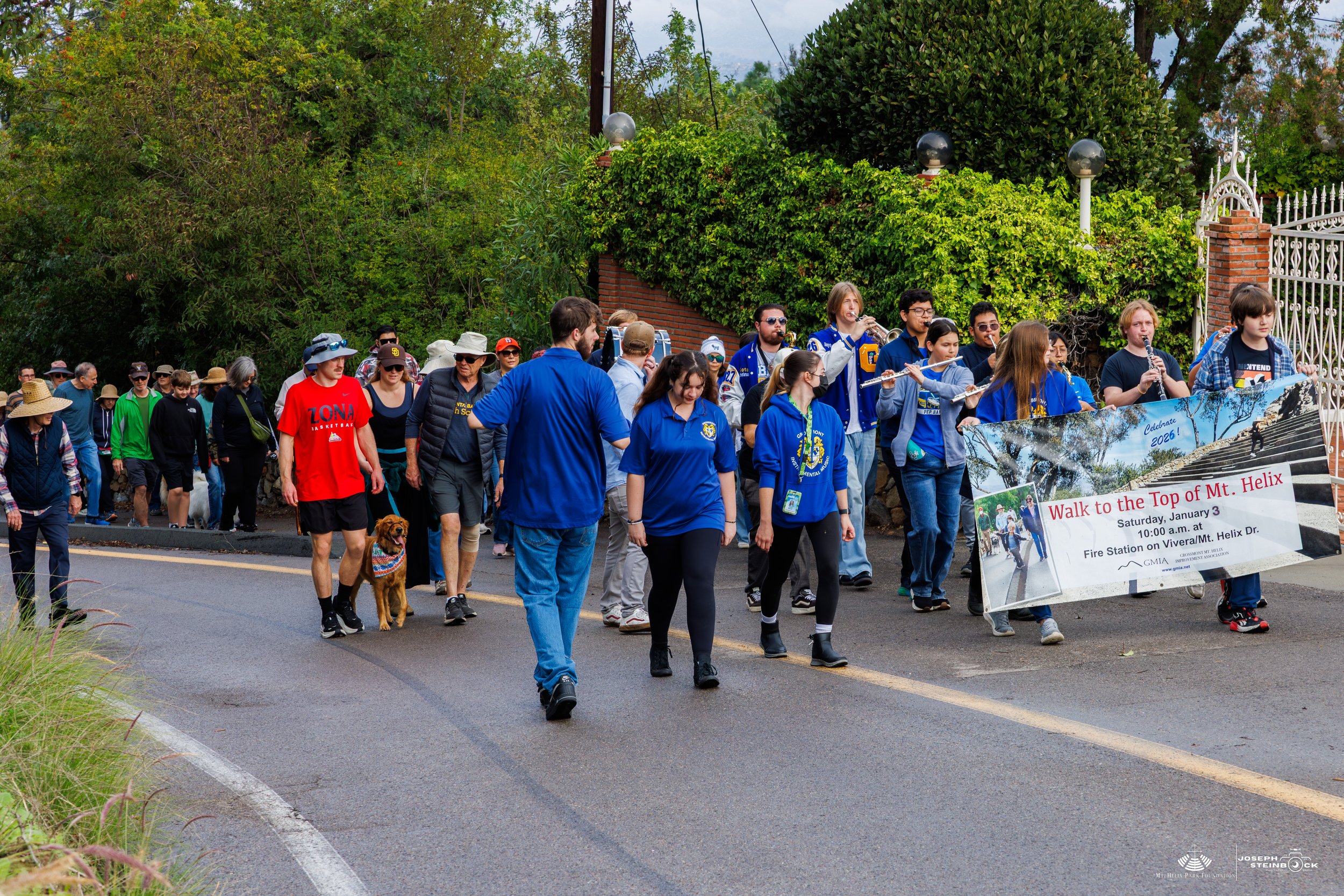 Group of people participating in a walk to the top of Mount Helix, some playing musical instruments, walking along a residential street with green trees and a decorative gate in the background.