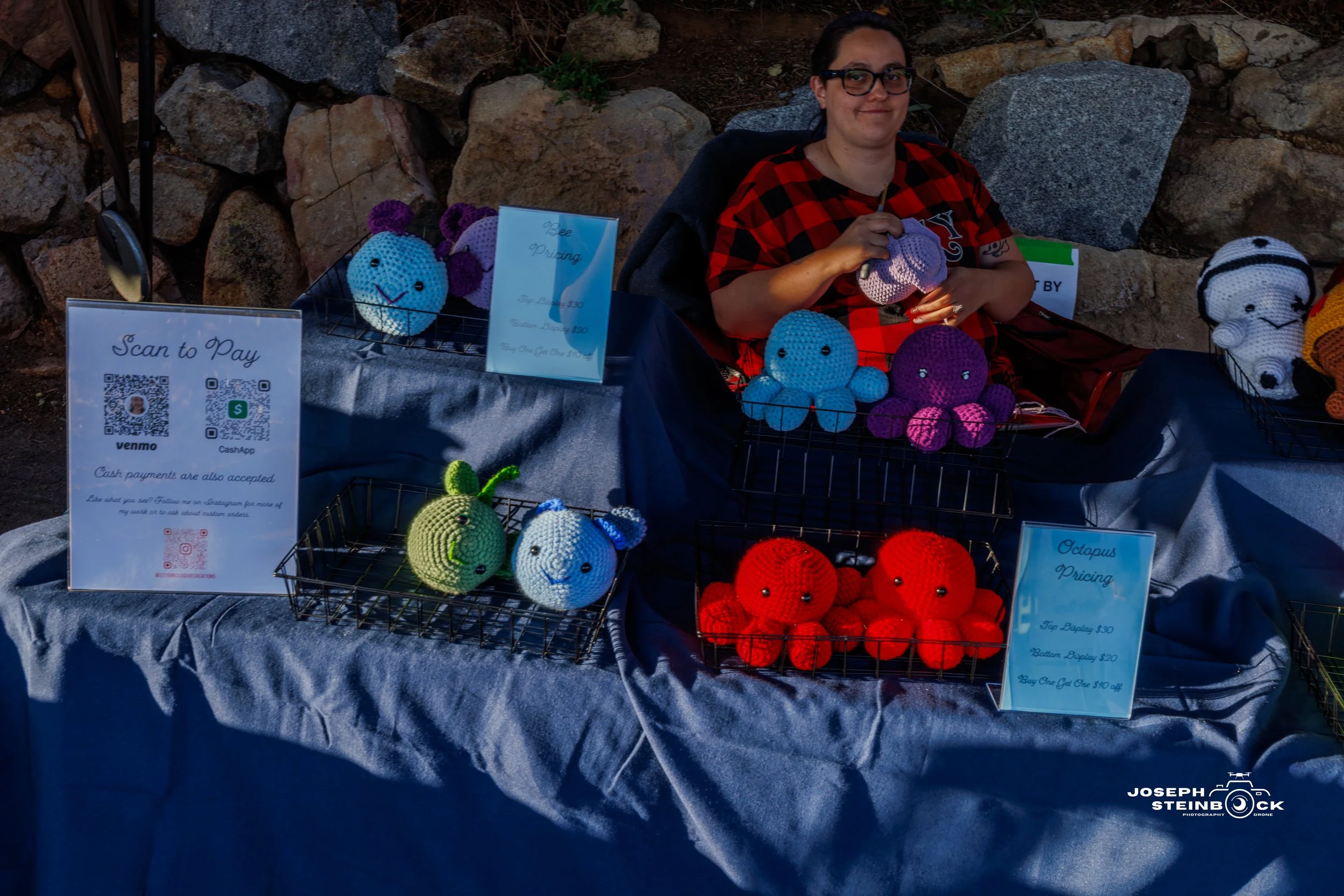 A woman sitting behind a table displaying crocheted octopus and bee toys at a craft sale. The table has signs with pricing and a QR code for digital payments. The background is a stone wall.
