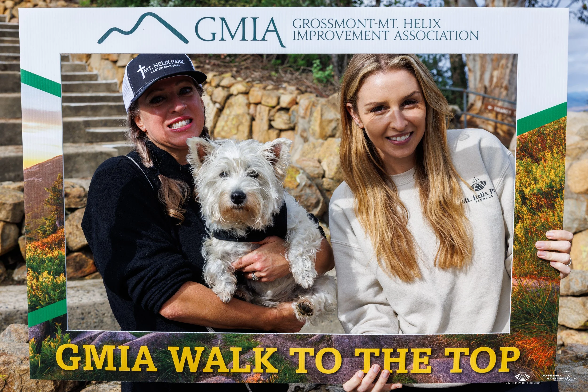 Two smiling women and a small white dog posing behind a sign that reads "GMIA Walk to the Top" at Mt. Helix. One woman is wearing a black cap and jacket, and the other is wearing a cream hoodie. The background shows stone stairs and rocks.