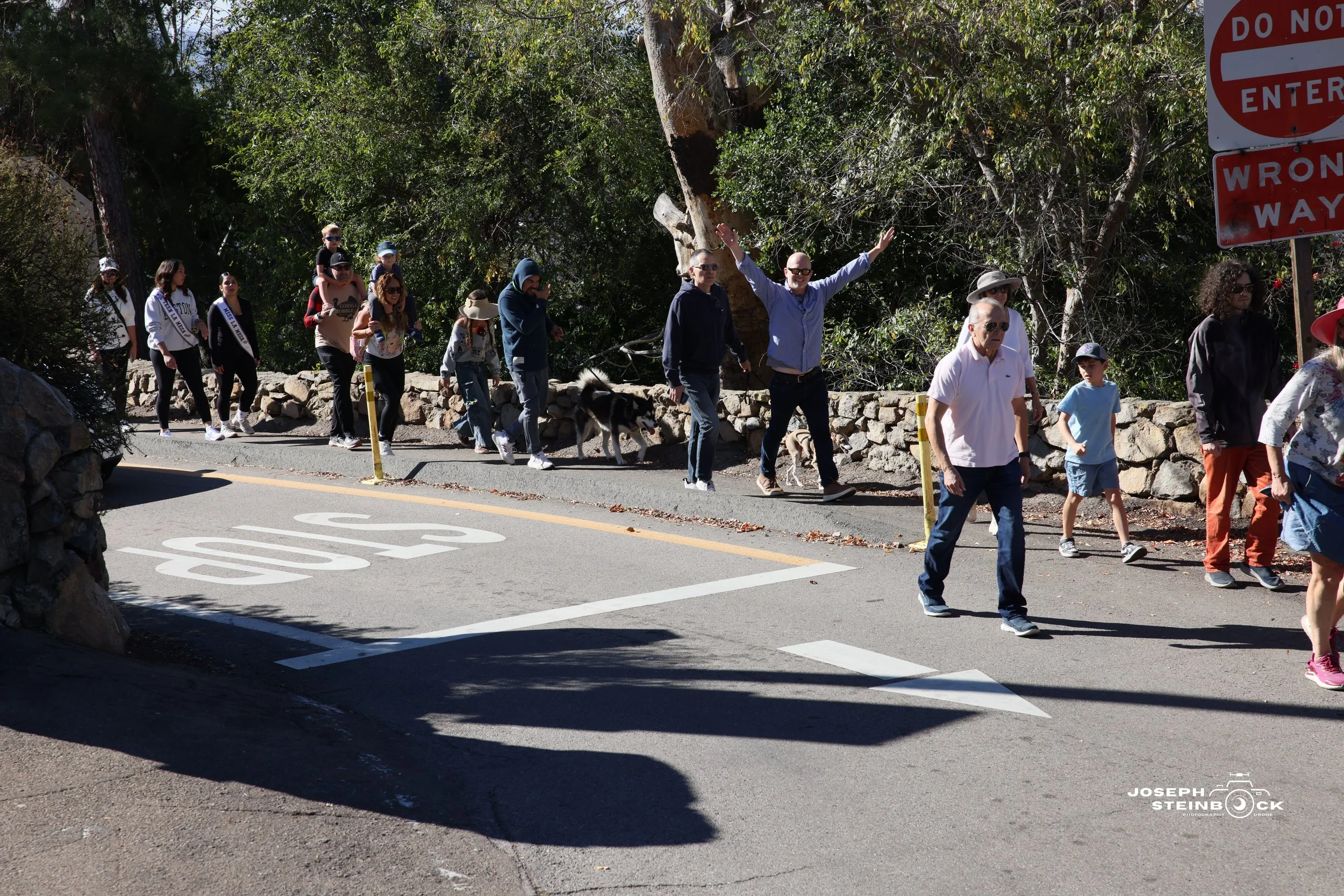 A group of people, including adults and children, walking on a sidewalk near a stone wall and trees, with some wearing sunglasses and hats. There is a large red and white 'Do Not Enter' sign and a painted stop sign on the street.