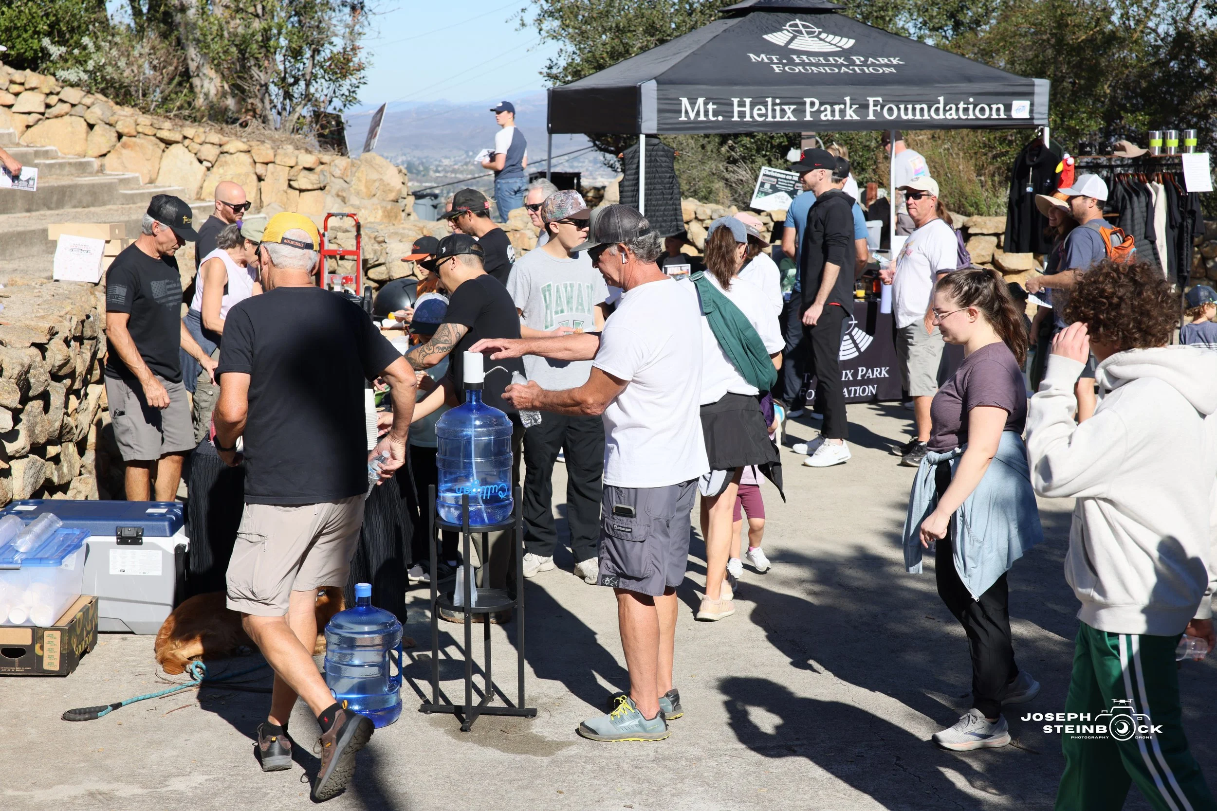 Group of people gathered at an outdoor event under a black canopy with the text 'Mt. Helix Park Foundation'. People are standing in line, talking, and enjoying the event in a sunny setting with trees and stone steps in the background.