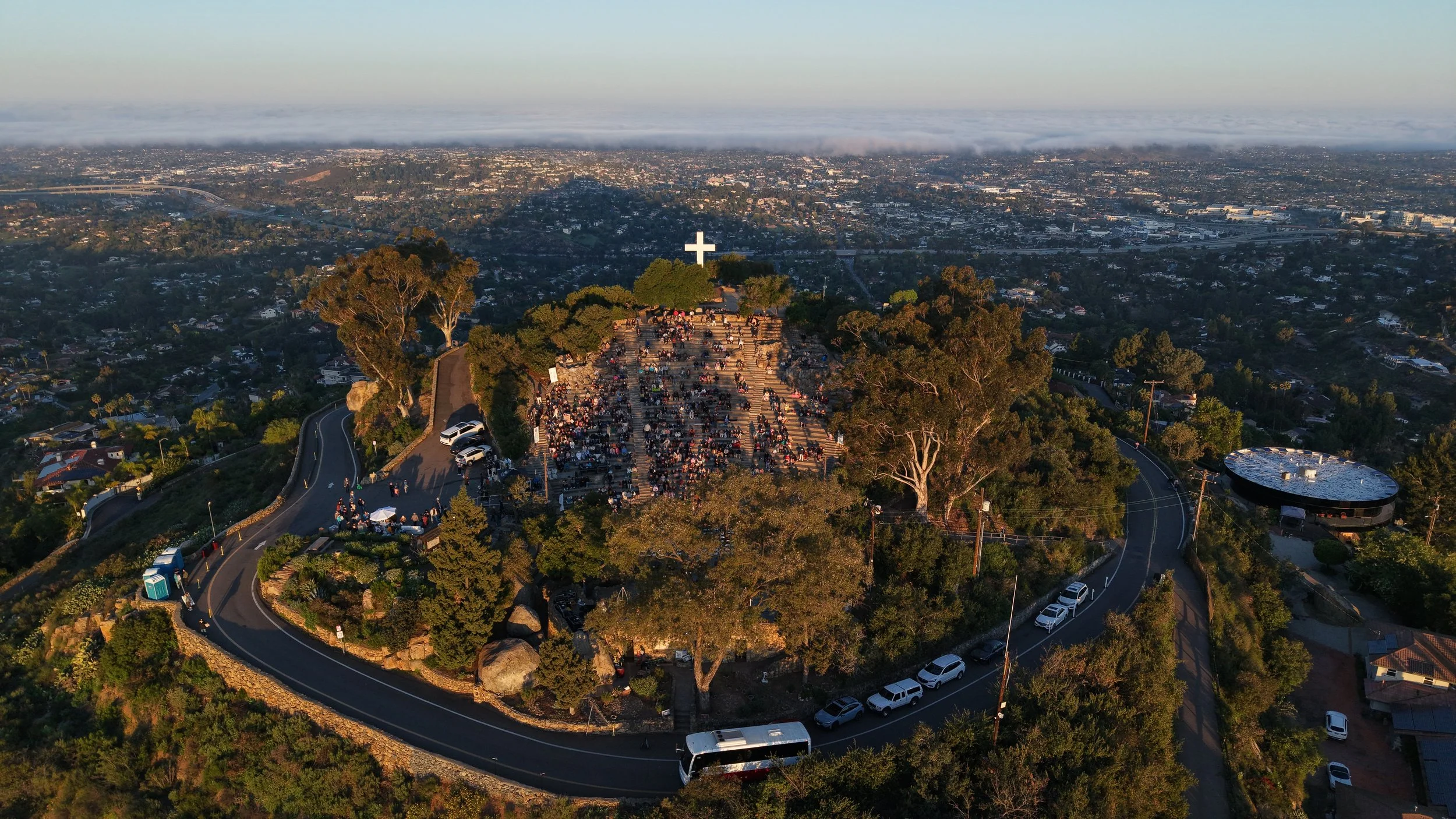 Aerial view of a hillside cemetery with a large cross at the top, filled with people attending a gathering, overlooking a cityscape in the background.