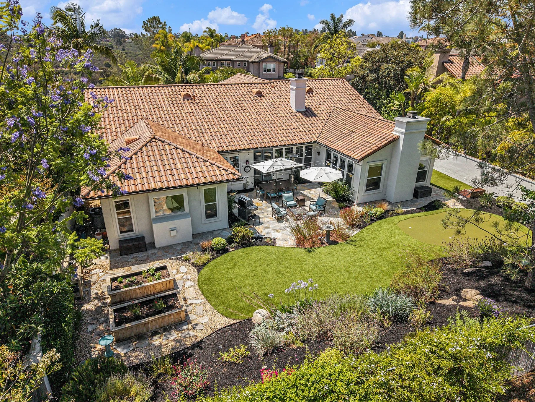 Aerial view of a backyard with a well-maintained lawn, flower beds, a stone pathway, and outdoor seating with umbrellas, adjacent to a house with a terracotta tiled roof surrounded by trees and neighboring houses in the background.