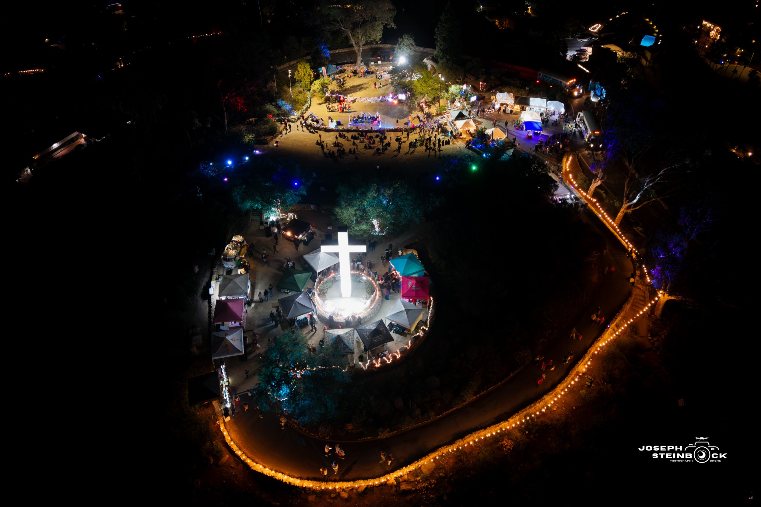 Night aerial view of a gathering at a park centered around a lit white cross monument, with tents, pathways decorated with string lights, and people attending the event.