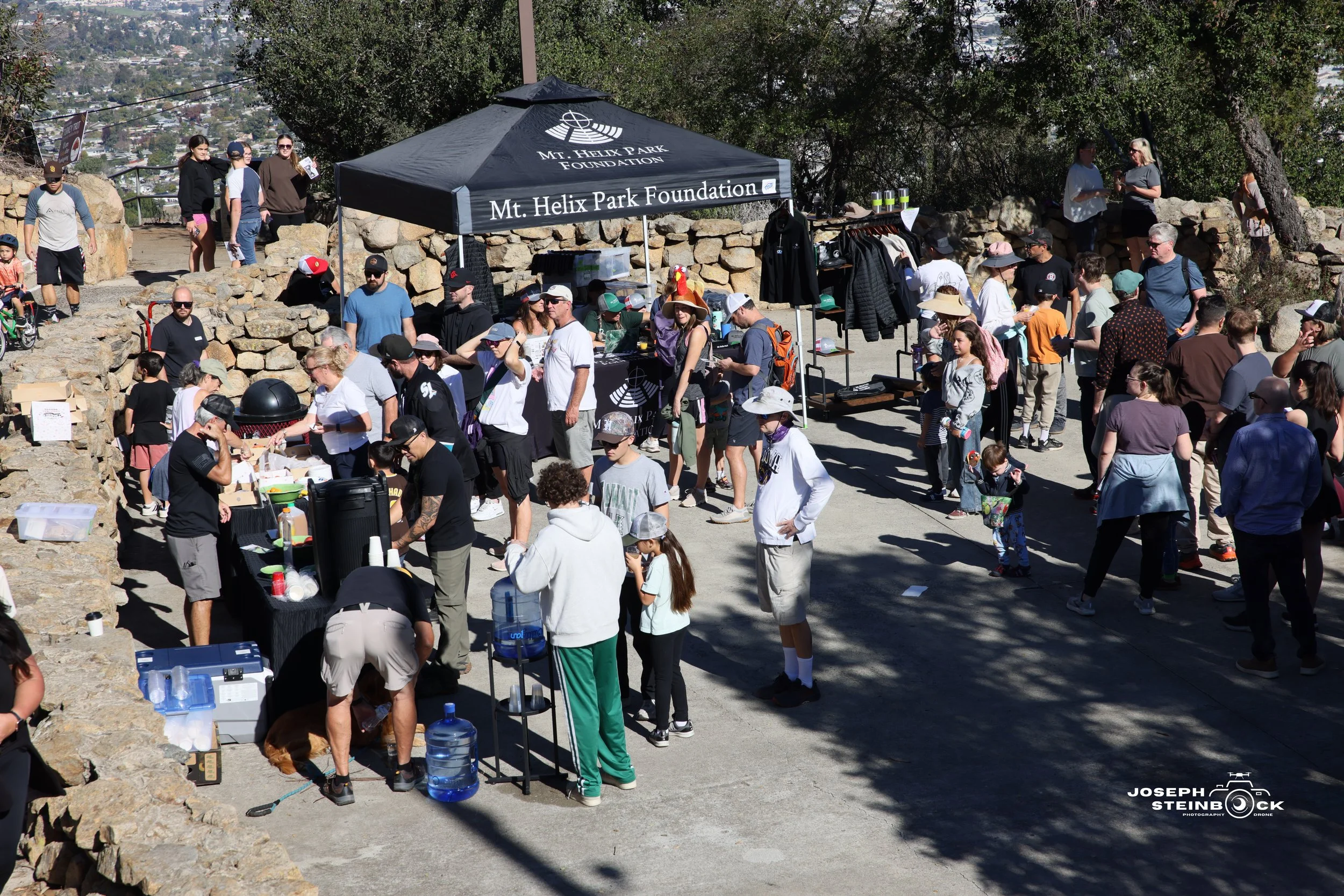 Crowd of people gathered at an outdoor event at Mt. Helix Park, with a black tent labeled 'Mt. Helix Park Foundation,' stone walls, and trees in the background.