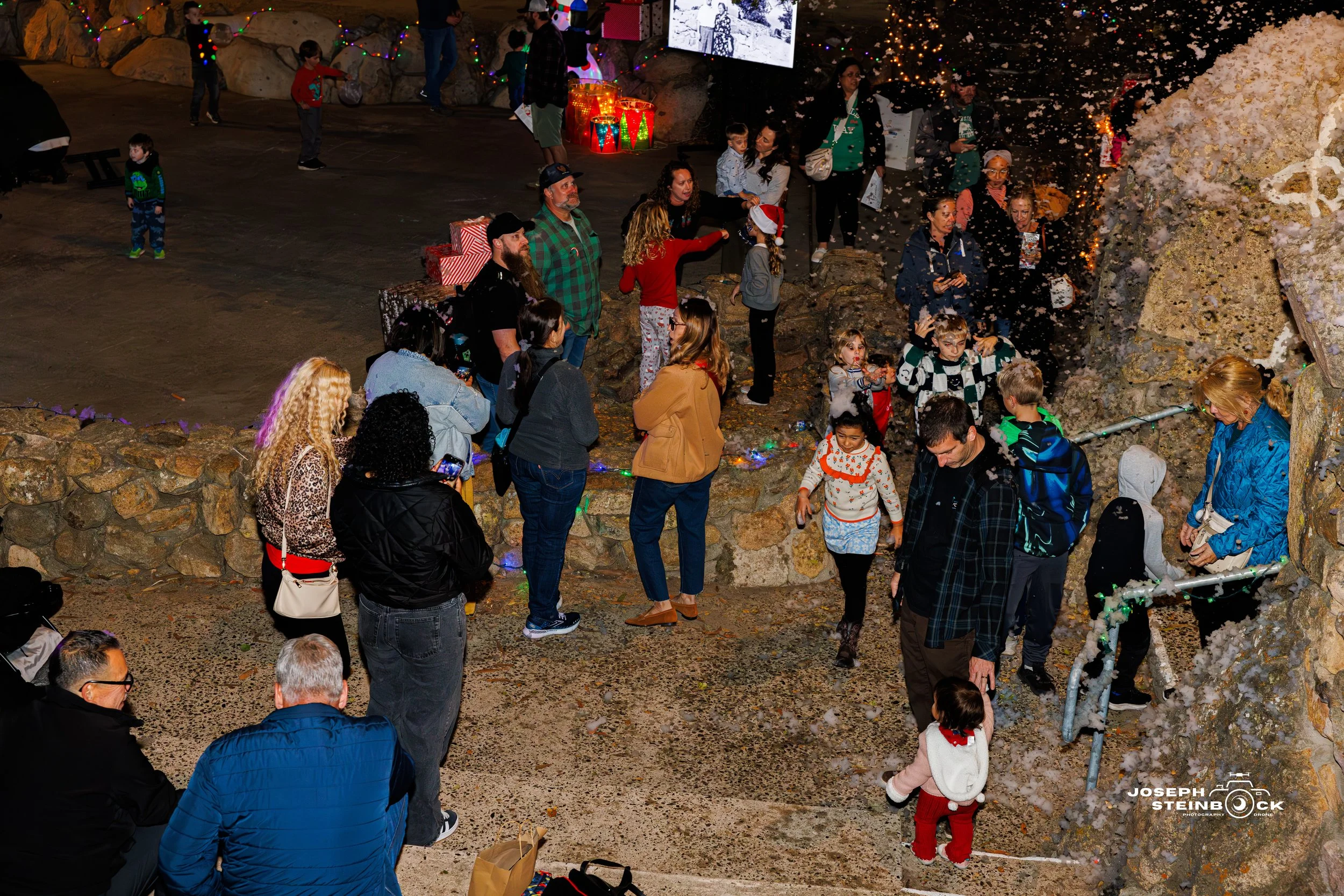 Children and adults gathering outdoors at night, some wearing festive holiday sweaters and Santa hats, with Christmas decorations and lights, as some throw snow or confetti.