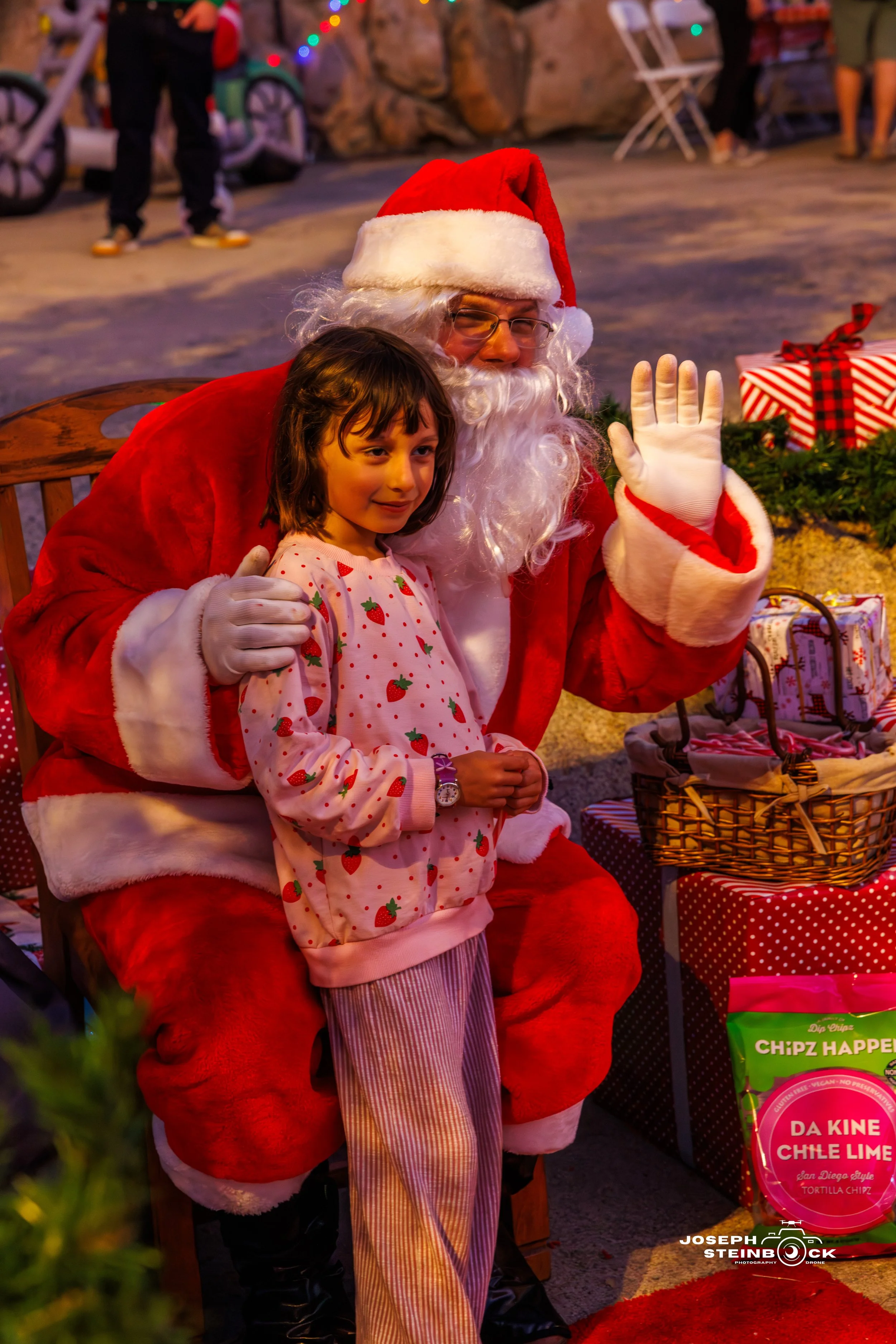 A young girl stands next to Santa Claus, who is dressed in a red suit with white fur trim, at a festive holiday event. Santa is waving and smiling, behind a table decorated with Christmas presents and greenery.