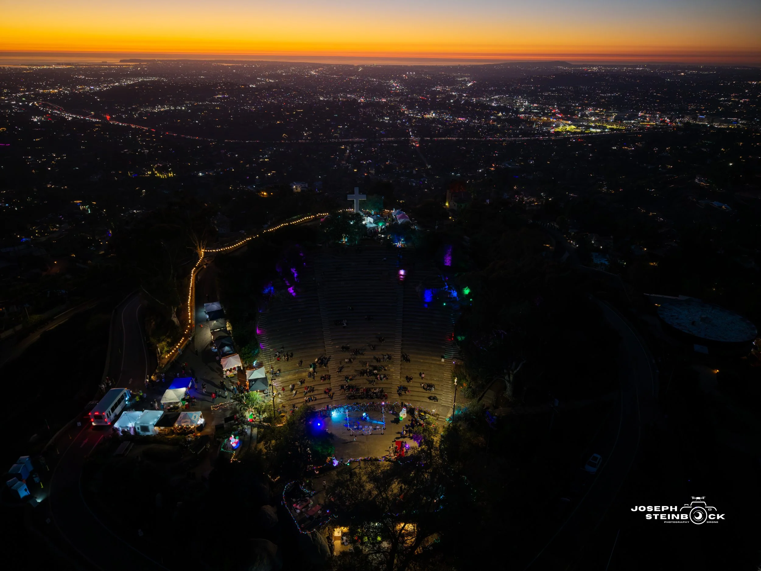 Aerial view of an outdoor event at dusk with a large crowd in an amphitheater, surrounded by colorful lights, trees, tents, and a cross at the top of a hill. City lights and a sunset horizon are visible in the distance.