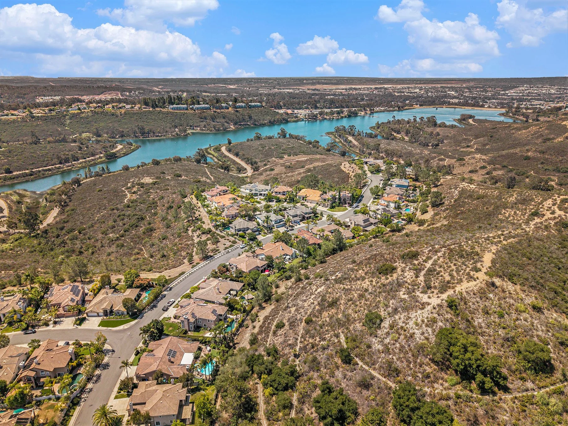 Aerial view of a suburban neighborhood with houses, winding roads, and a river in the background, surrounded by dry hills and green trees.