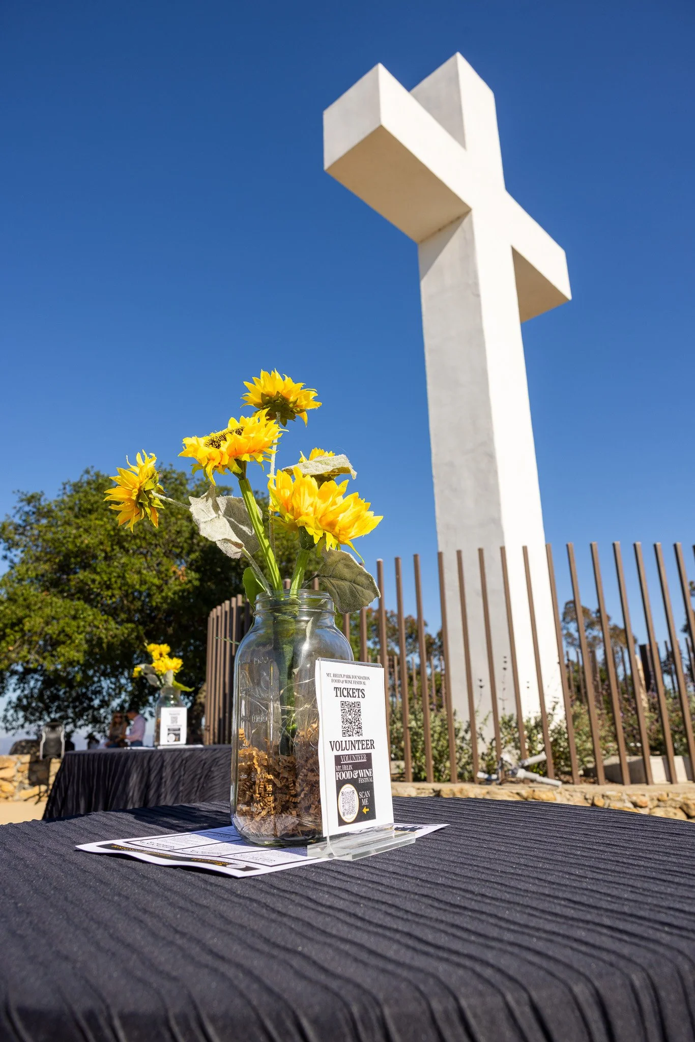 Yellow sunflower bouquet in a glass jar with a ticket stand on a black tablecloth, set against a large white cross monument and a clear blue sky.