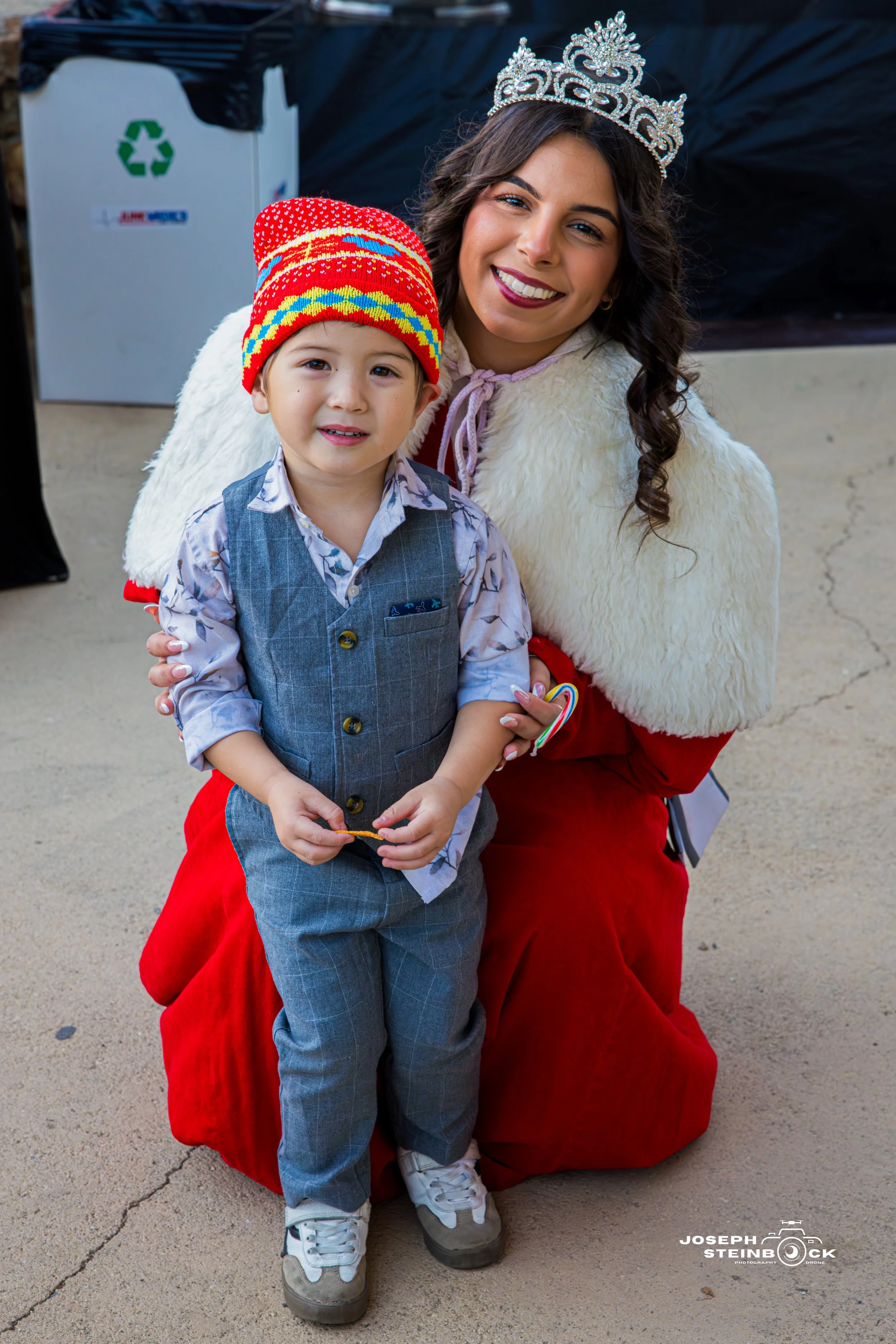 A woman dressed as Santa Claus wearing a crown, smiling and posing with a young boy in festive clothing and a colorful knitted hat.
