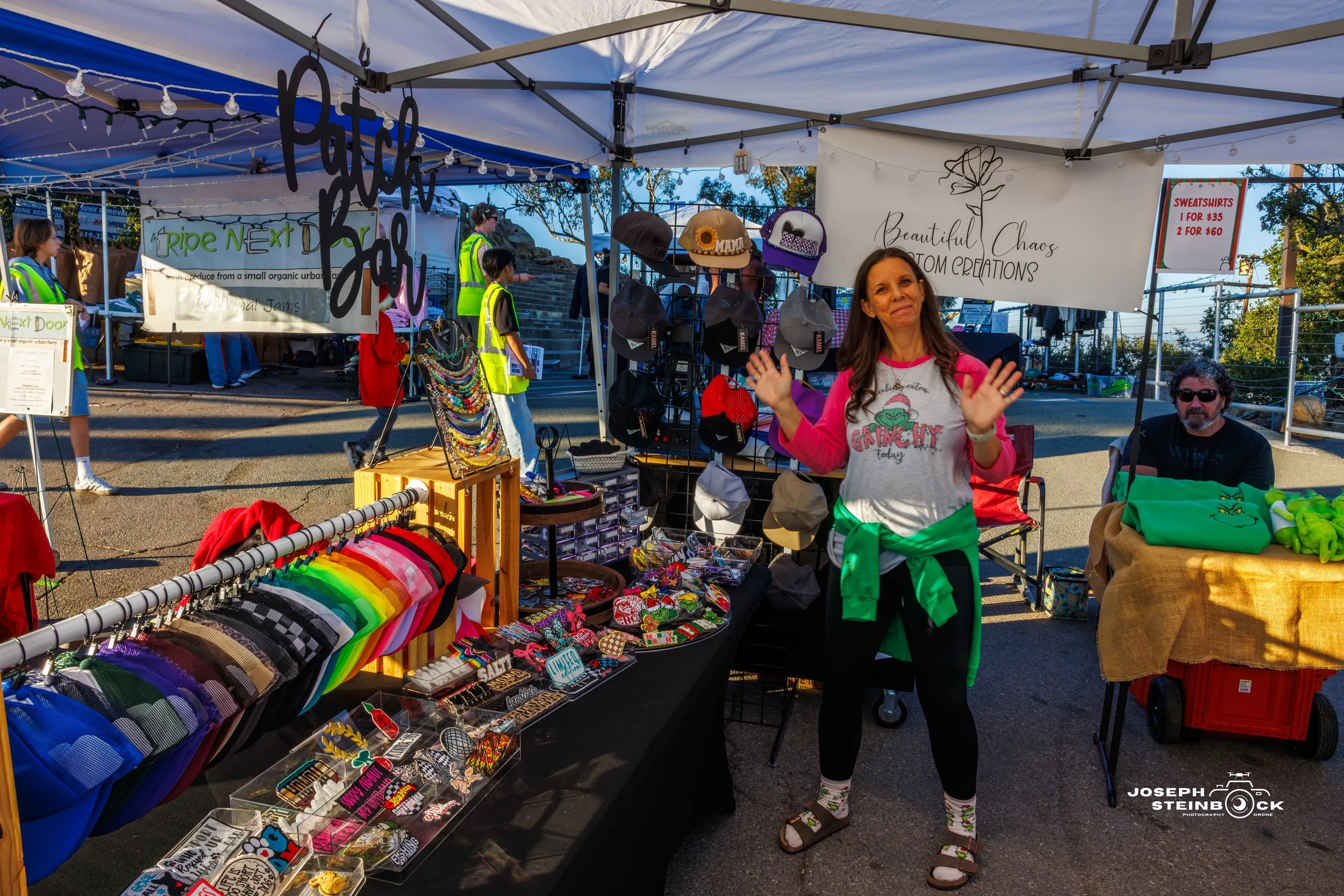 A woman with long brown hair wearing a white shirt with pink and grey sleeves, black leggings, sandals, and socks is standing at an outdoor market booth. She is smiling and waving with both hands. The booth displays colorful hats, bracelets, patches,