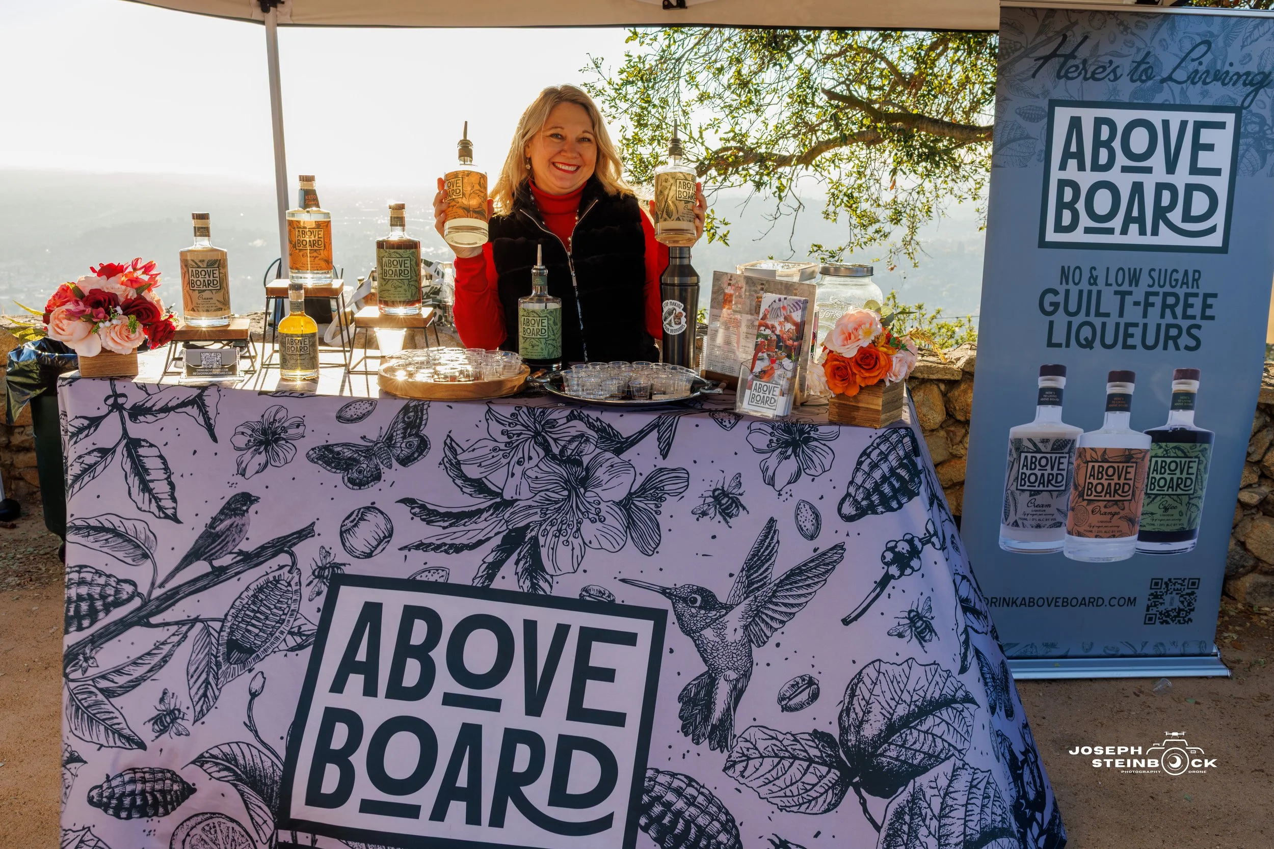 A cheerful woman standing behind a table displaying bottles of Above Board liqueur, set up outdoors under a tent with scenic mountain views in the background. The table has a floral-patterned tablecloth and a banner with the Above Board logo, promoti