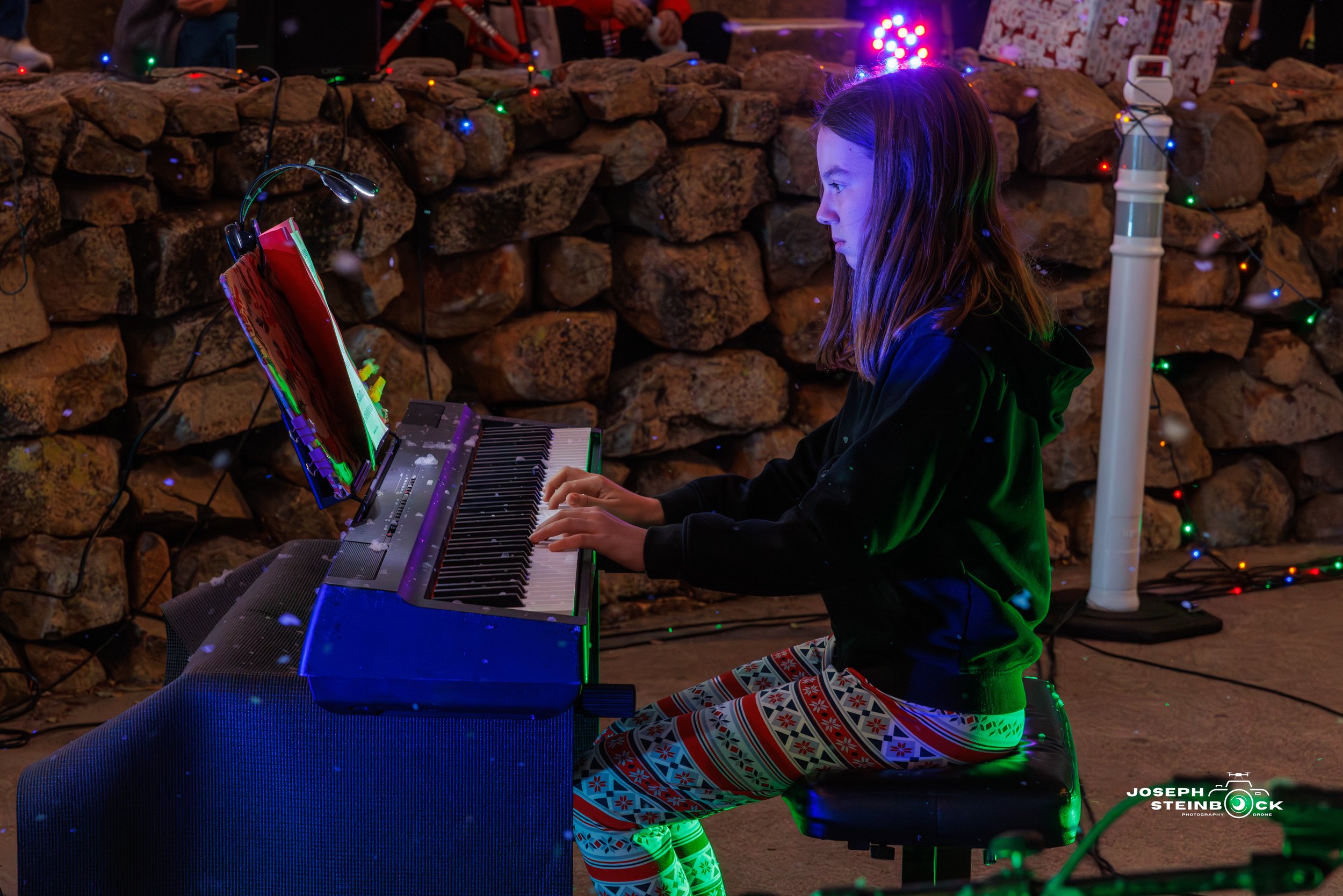 A young girl playing a digital piano during a holiday celebration decorated with colorful string lights. She is wearing festive pajamas with red, white, and blue patterns and has long brown hair. The background features a stone wall, and a subtropica