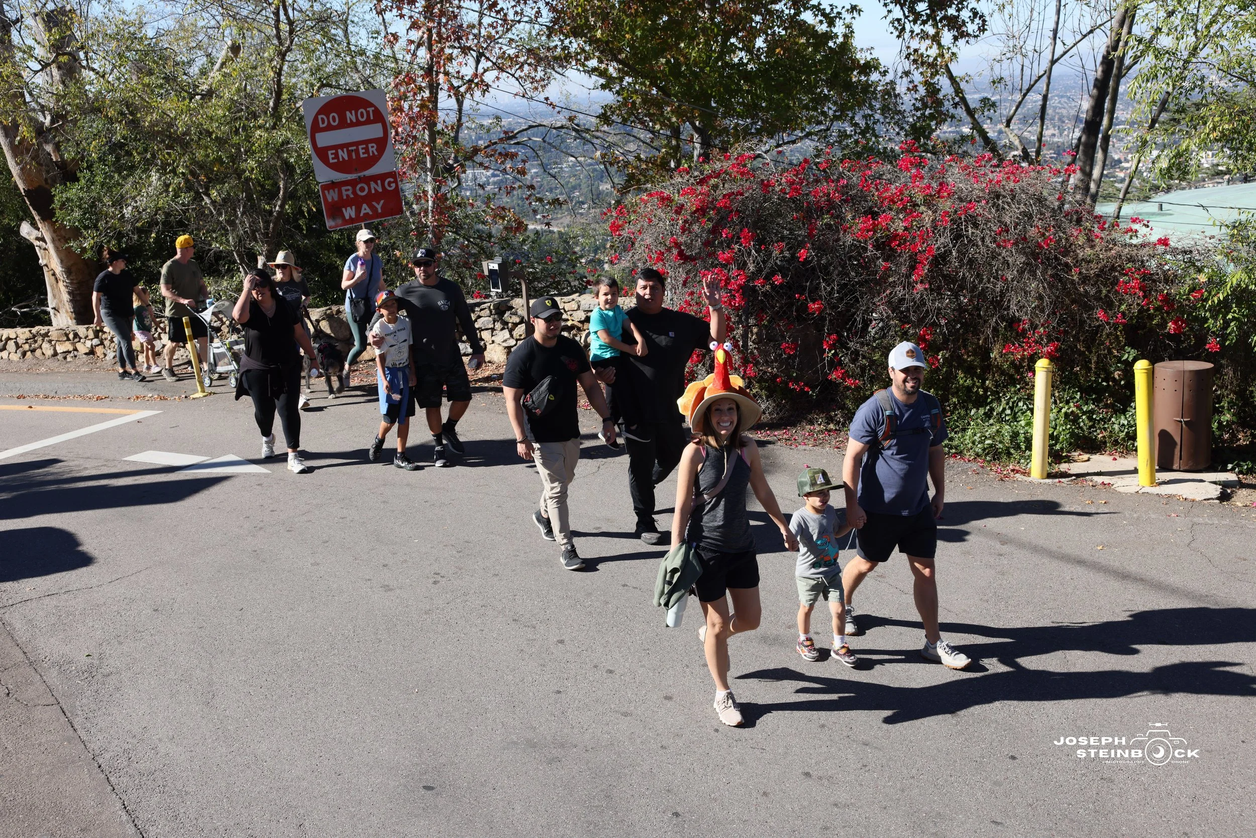 A group of people walking outdoors on a paved path in a park or nature area, with some wearing hats and casual clothing. The background has trees, bushes with red flowers, and a 'Do Not Enter, Wrong Way' sign. The people appear to be enjoying a leisu