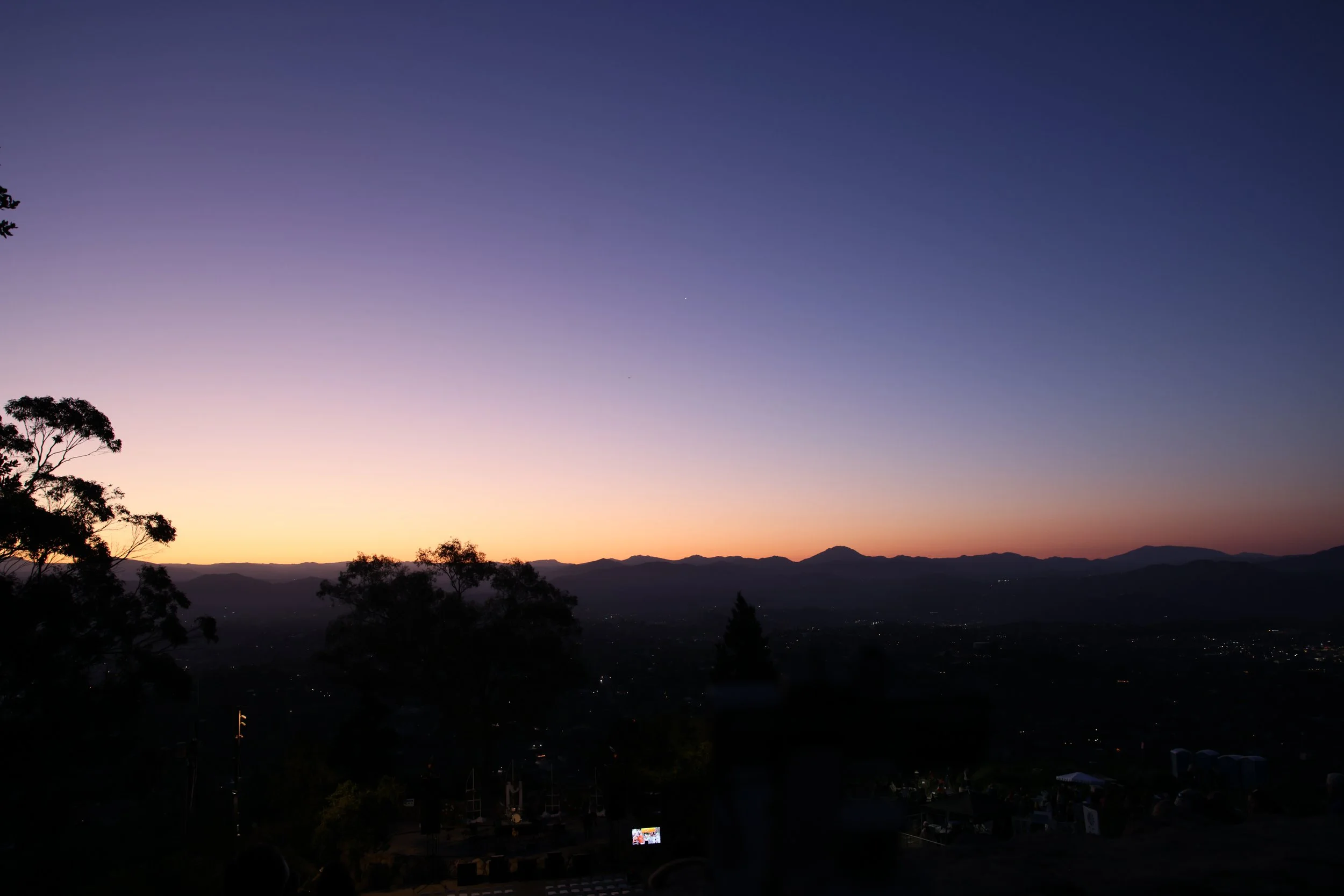 Sunset or dawn over a mountainous landscape with silhouettes of trees and city lights below.