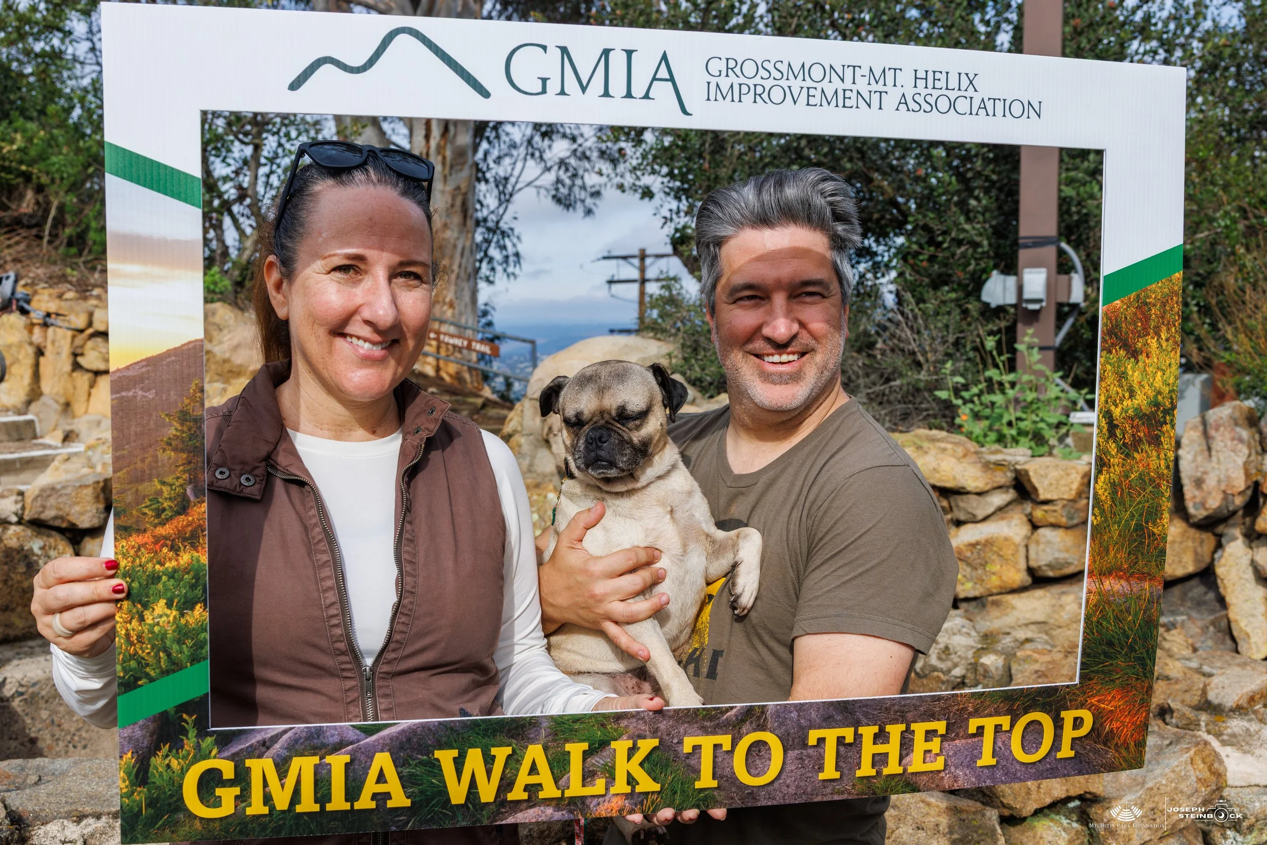 A smiling woman and man holding a small dog inside a frame at the GMIA Walk to the Top event, with a rocky outdoor landscape behind them.