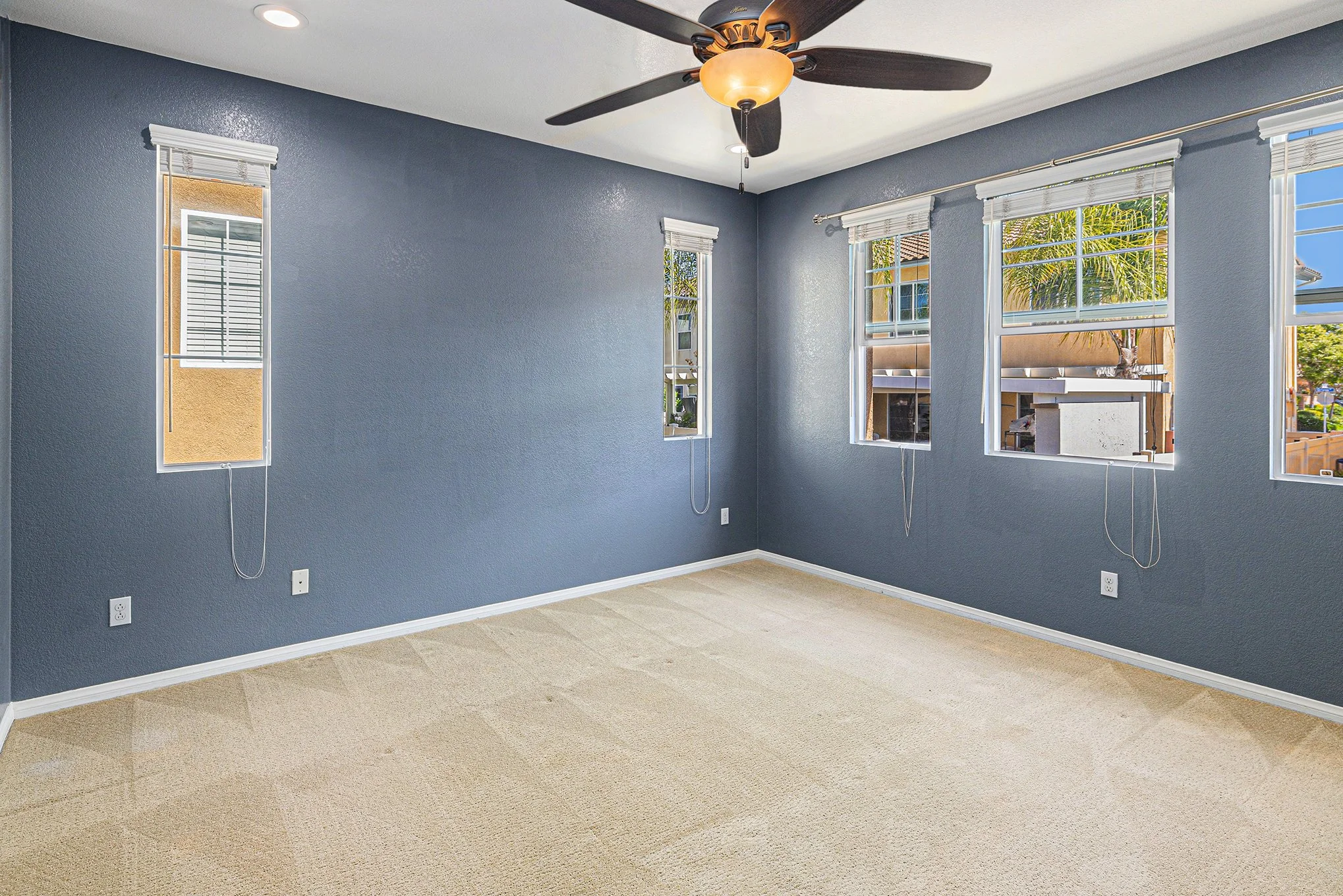 Empty room with blue walls, beige carpet, four windows with blinds, a ceiling fan with light, and outdoor view of palm trees.