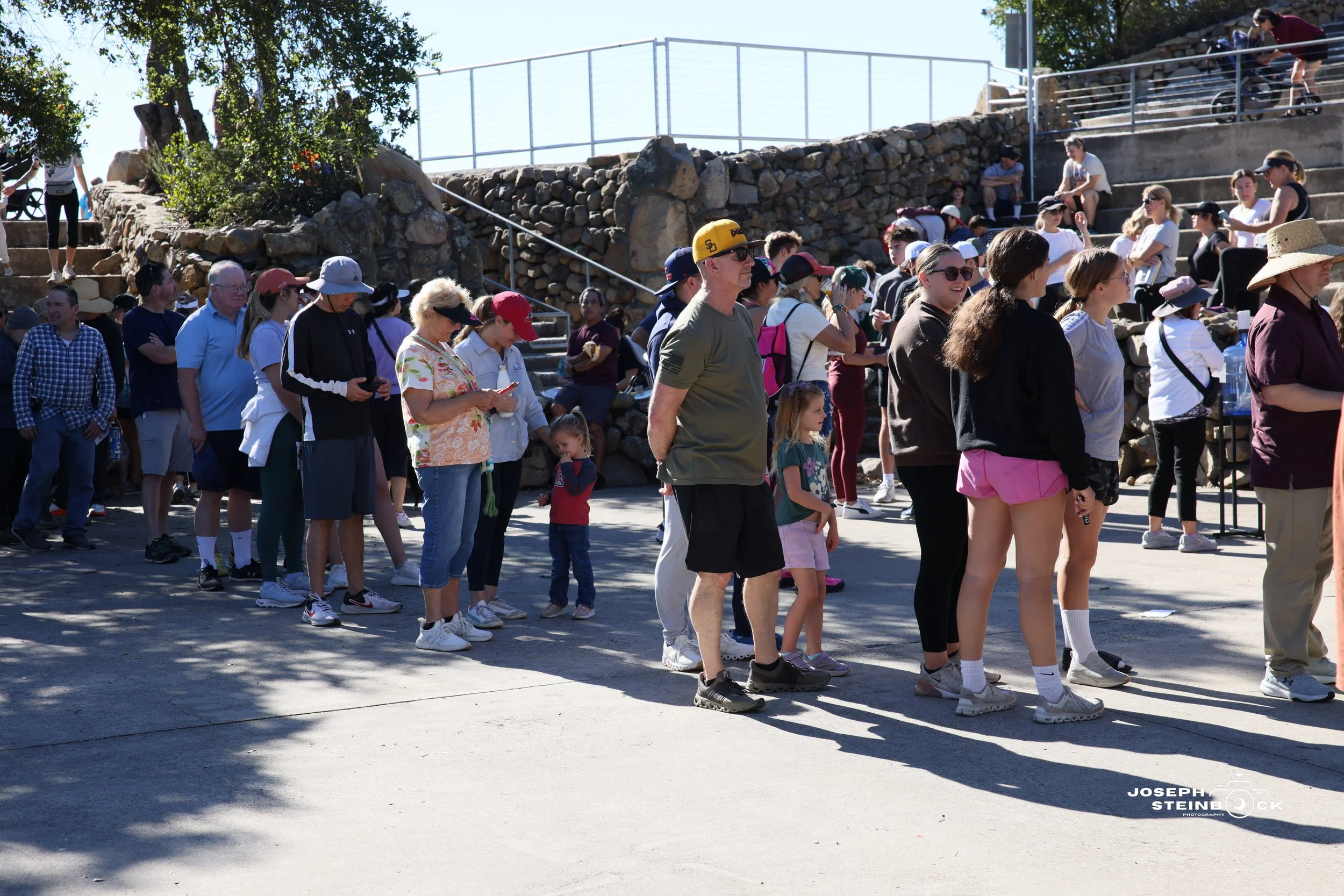 People standing in line outdoors on a sunny day, with some sitting on stairs and others on the ground, in front of a stone wall and railing.