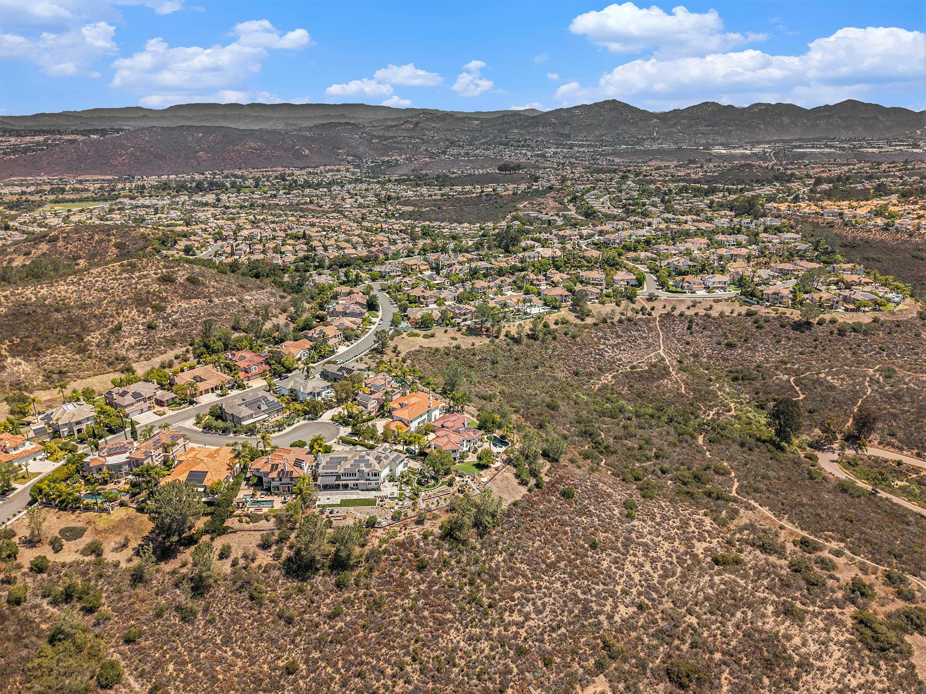 Aerial view of a suburban neighborhood surrounded by hilly, semi-arid terrain with sparse greenery and hiking trails, under a partly cloudy sky.