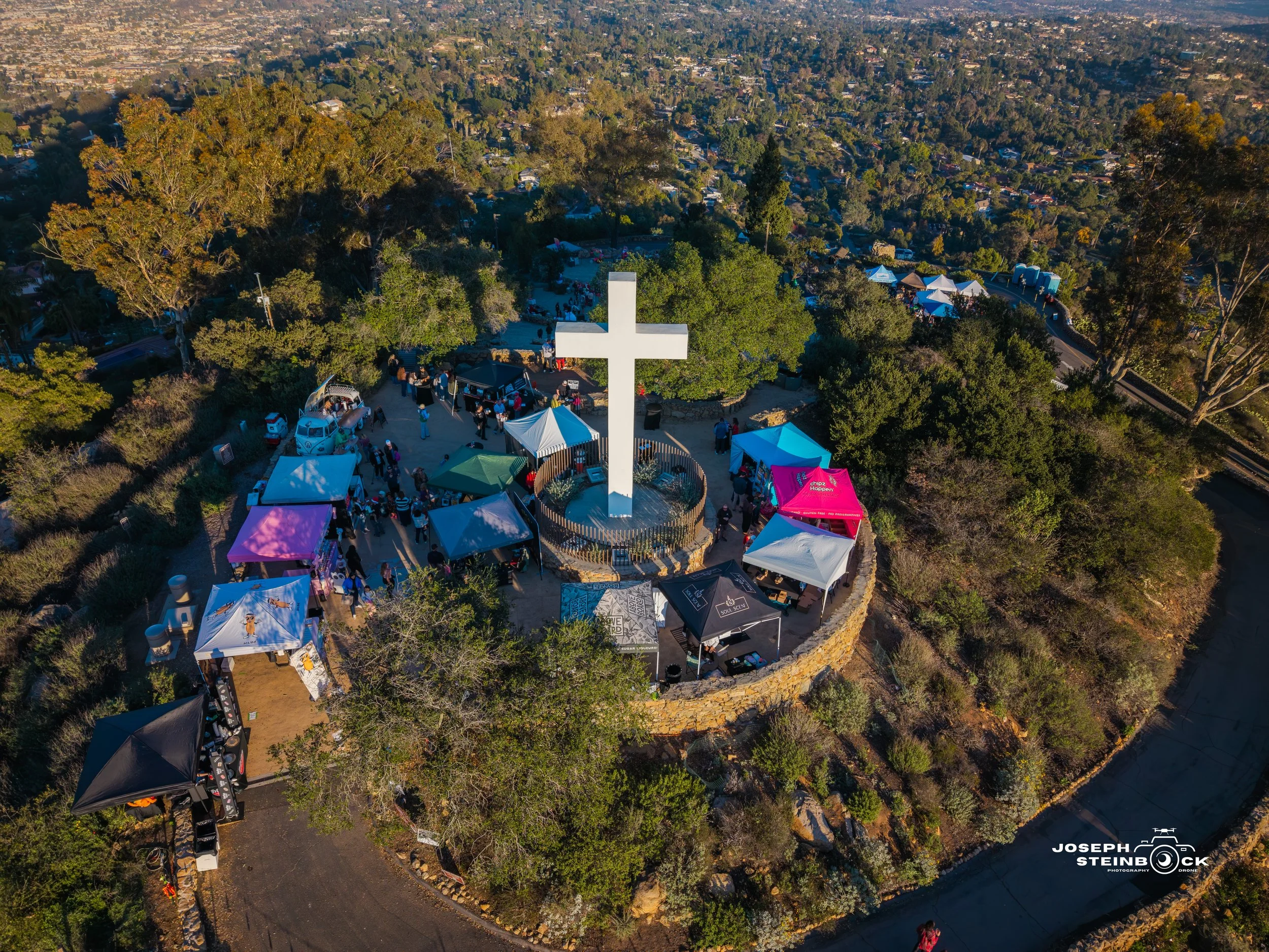 An aerial view of a large outdoor gathering at a park with a prominent white cross monument at the center, surrounded by trees, tents, and people.