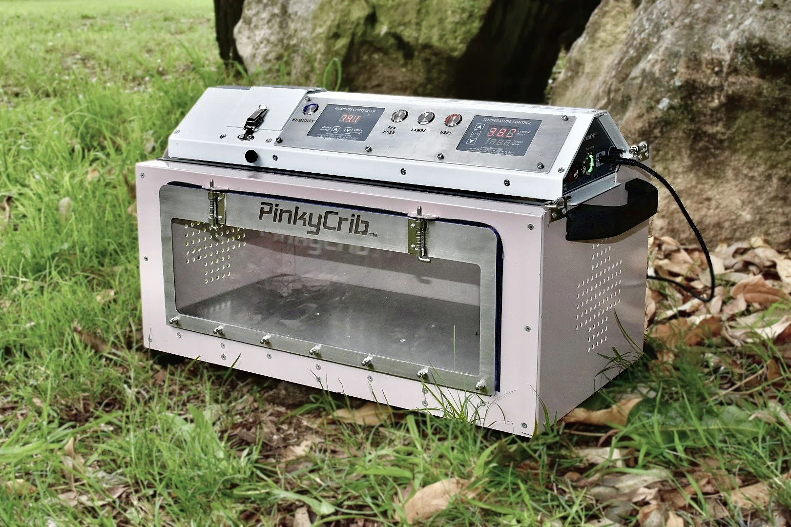 photo of PinkyCrib, a portable and compact wildlife intensive care unit on the grass with the control panel attached. Metallic frame around reflective surface with the text PinkyCrib