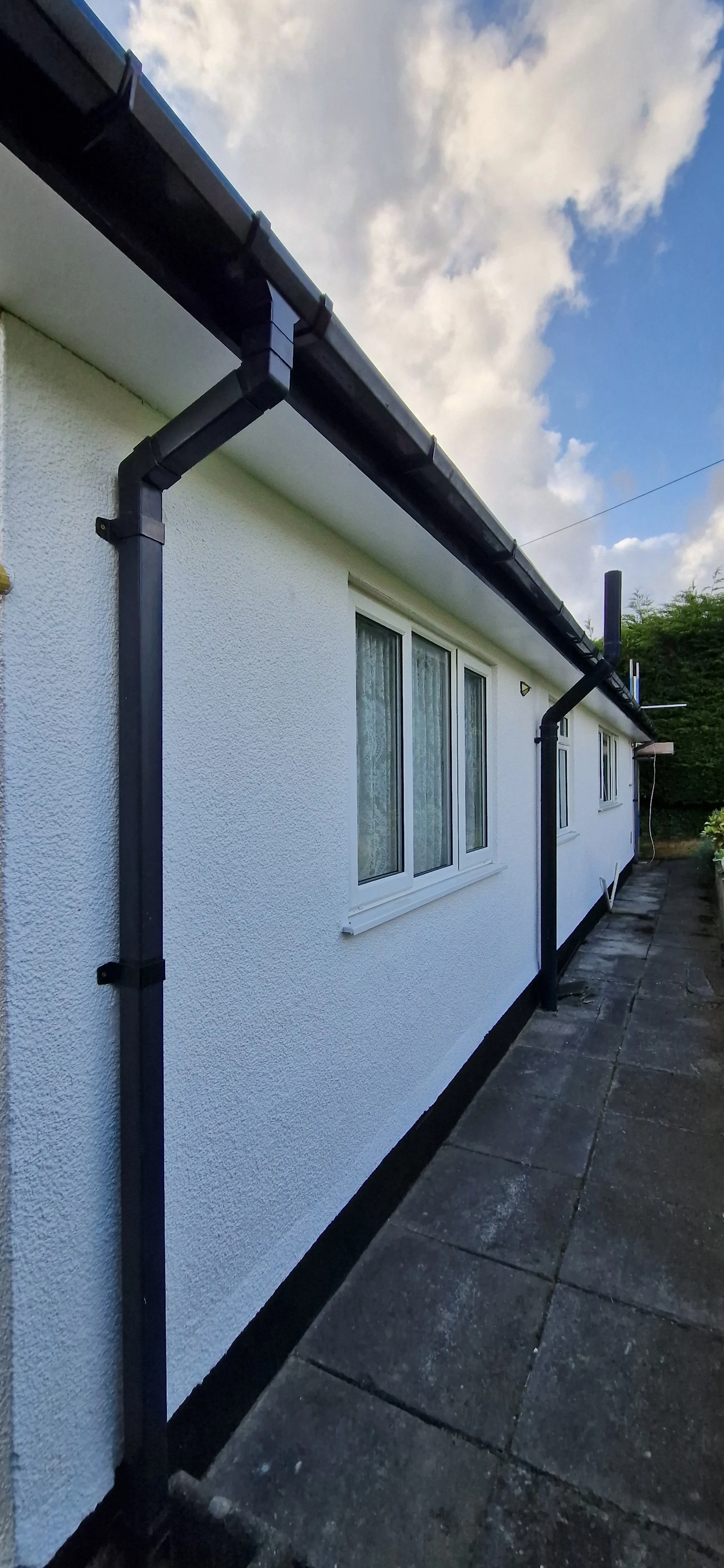 Side of a house with white textured exterior walls, three windows with lace curtains, black gutters, and a concrete walkway.