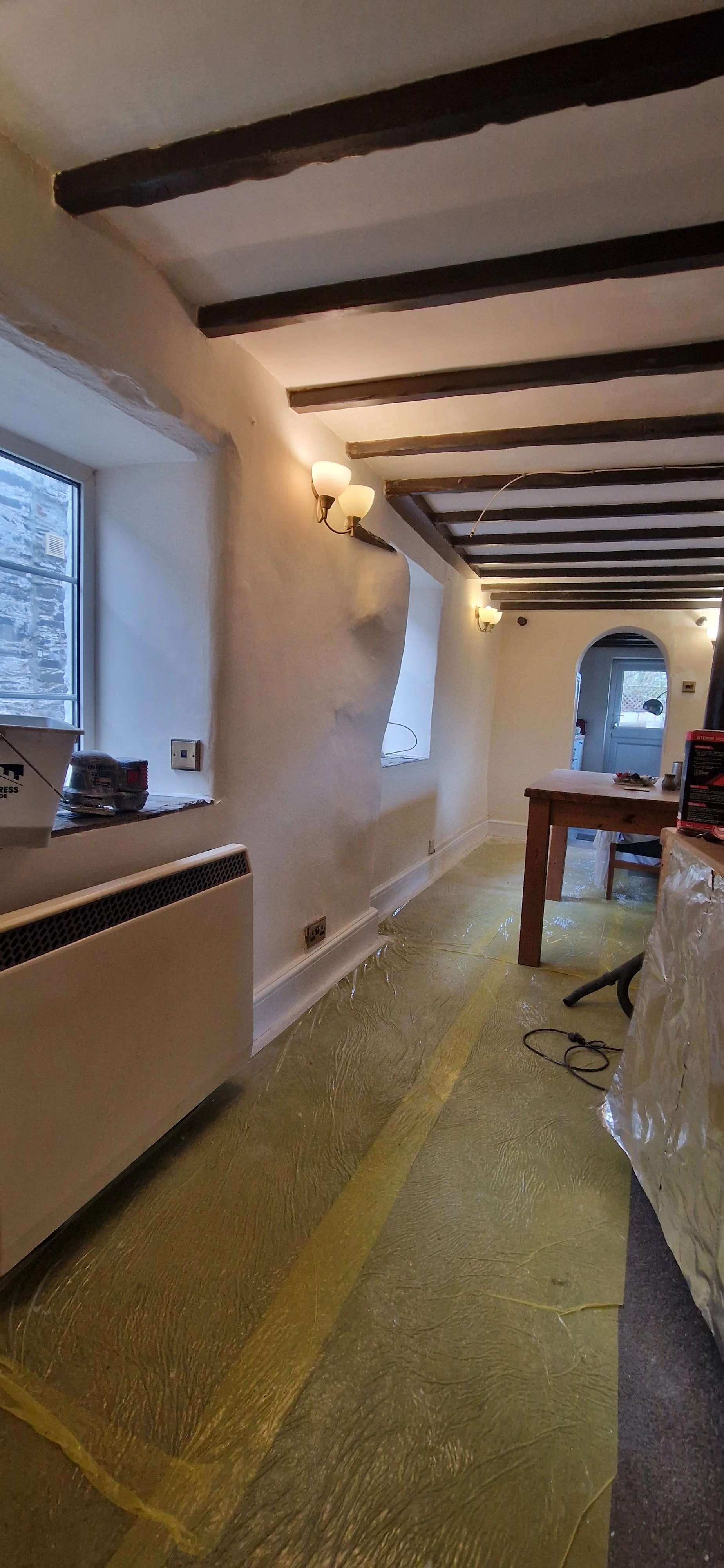 A room under renovation with plastic covering the wooden floor, a window on the left wall, dark wooden beams on the ceiling, and wall-mounted lights. Construction tools and materials are visible.