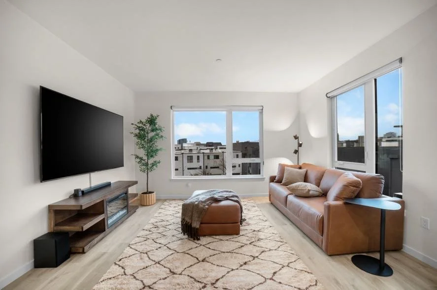 Bright living room with a large wall-mounted TV, a leather sofa with pillows, a matching ottoman with a throw blanket, a wooden entertainment unit, a potted plant, a tall floor lamp, a side table, a patterned rug, and two large windows showing an urb