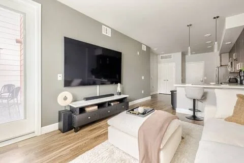 Living room with a mounted flat-screen TV, a white sofa with a beige blanket, a black TV stand, and an open kitchen in the background.