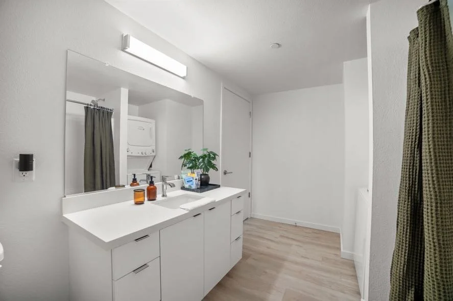 A modern bathroom with a large mirror, white countertop, and white cabinets. There are two amber-colored bottles on the counter, a green potted plant, and a few items near the plant. The room has white walls, a wooden floor, and a gray door. A dark g