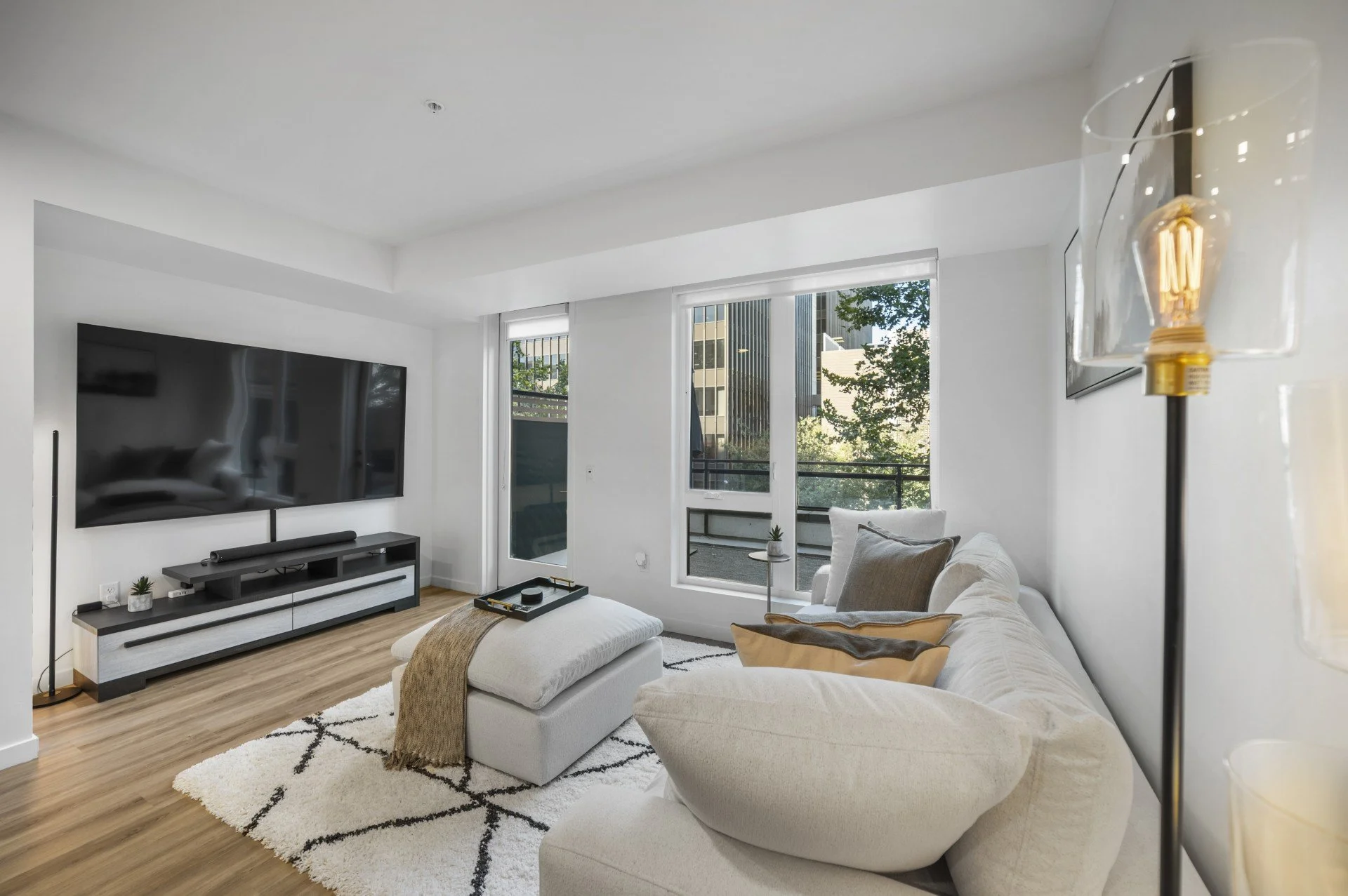 Living room with large flat-screen TV, white sofa with accent pillows, beige ottoman with a throw blanket, wooden floor, and large windows showing trees and buildings outside.