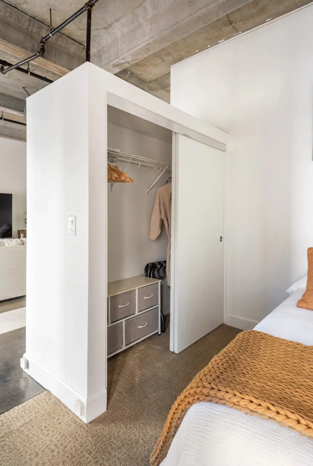Small bedroom with a white open closet containing a few hanging clothes and a beige storage box, next to a bed with white bedding and a chunky orange blanket.