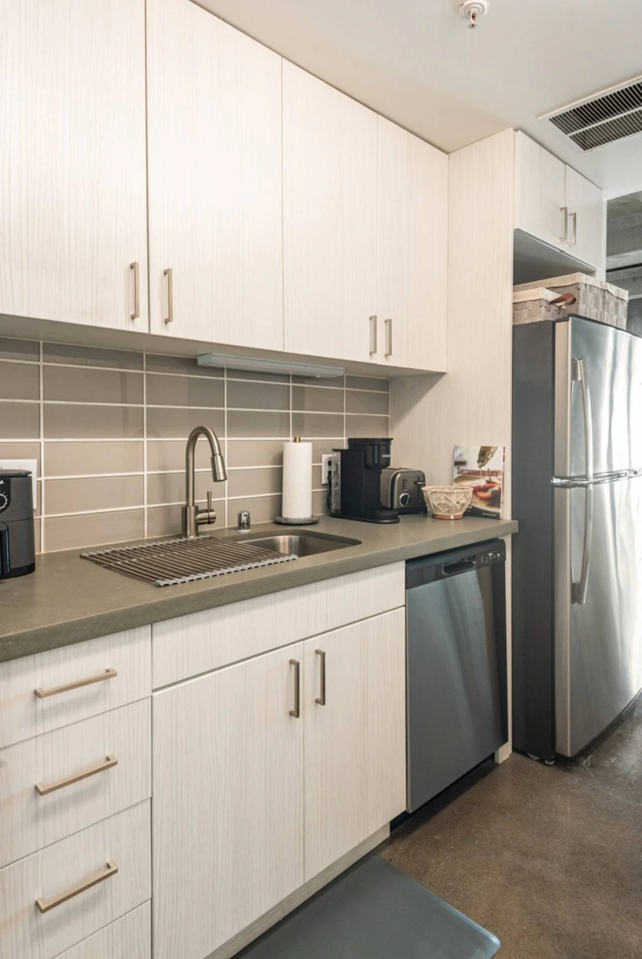 Kitchen with white cabinets, gray countertop, small sink, stainless steel refrigerator, and coffee maker.