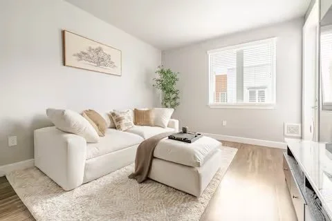 Living room with a white sofa, beige pillows, a tray with items on the ottoman, a beige blanket, a fluffy rug, a potted plant, and a window with blinds in a bright, minimalist space.