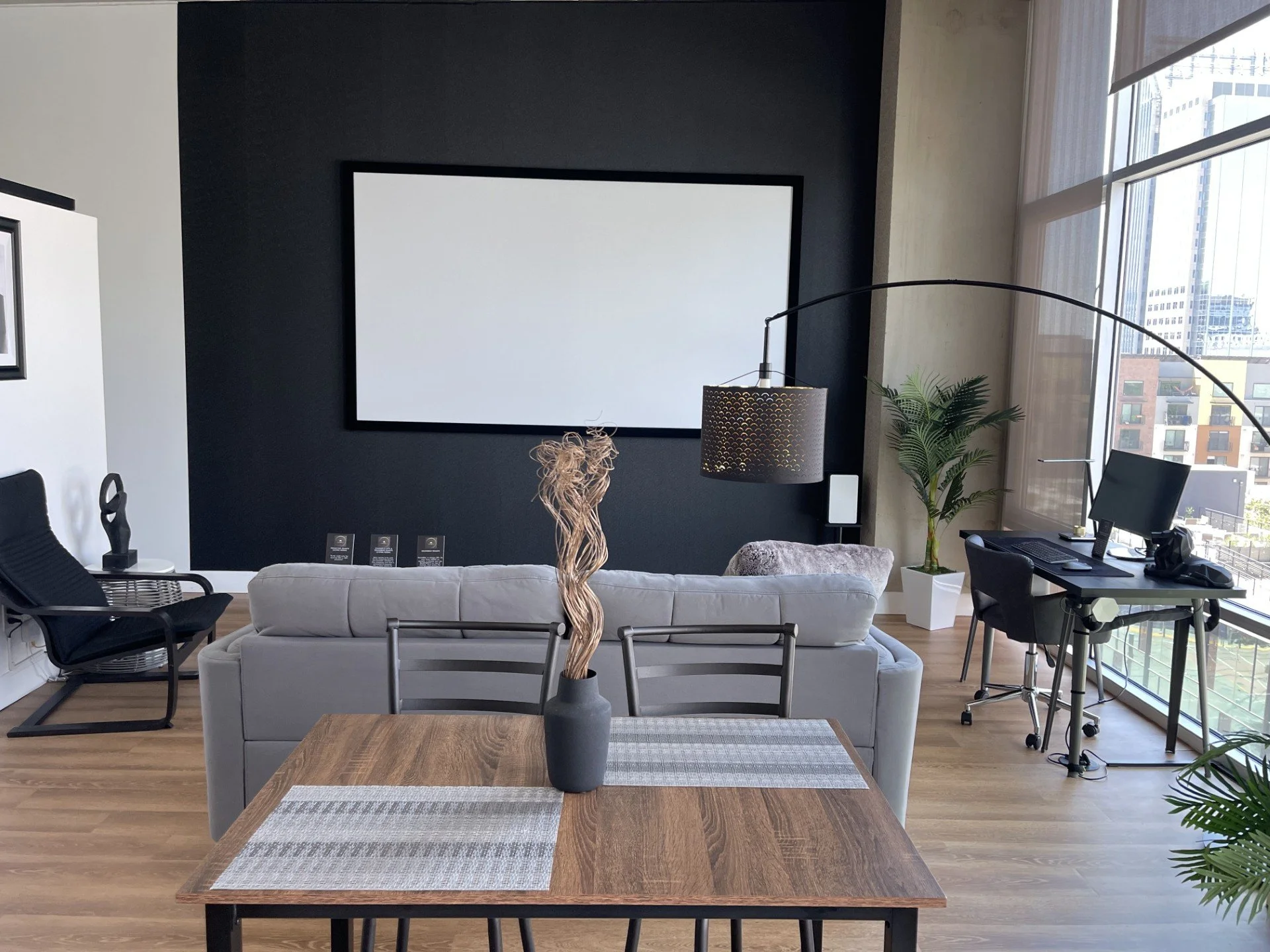 Modern office living room with a gray sofa, a wooden dining table with a black vase and decorative twigs, a black armchair, a large floor lamp, a potted plant, and a desk with a computer near large windows showing a city view.