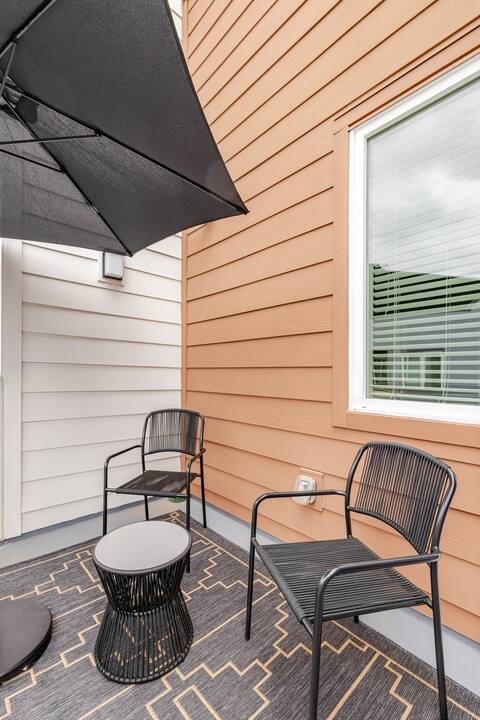 Patio with two black metal chairs, a black side table, a large black patio umbrella, beige and white siding, a window with white blinds, and a patterned outdoor rug.