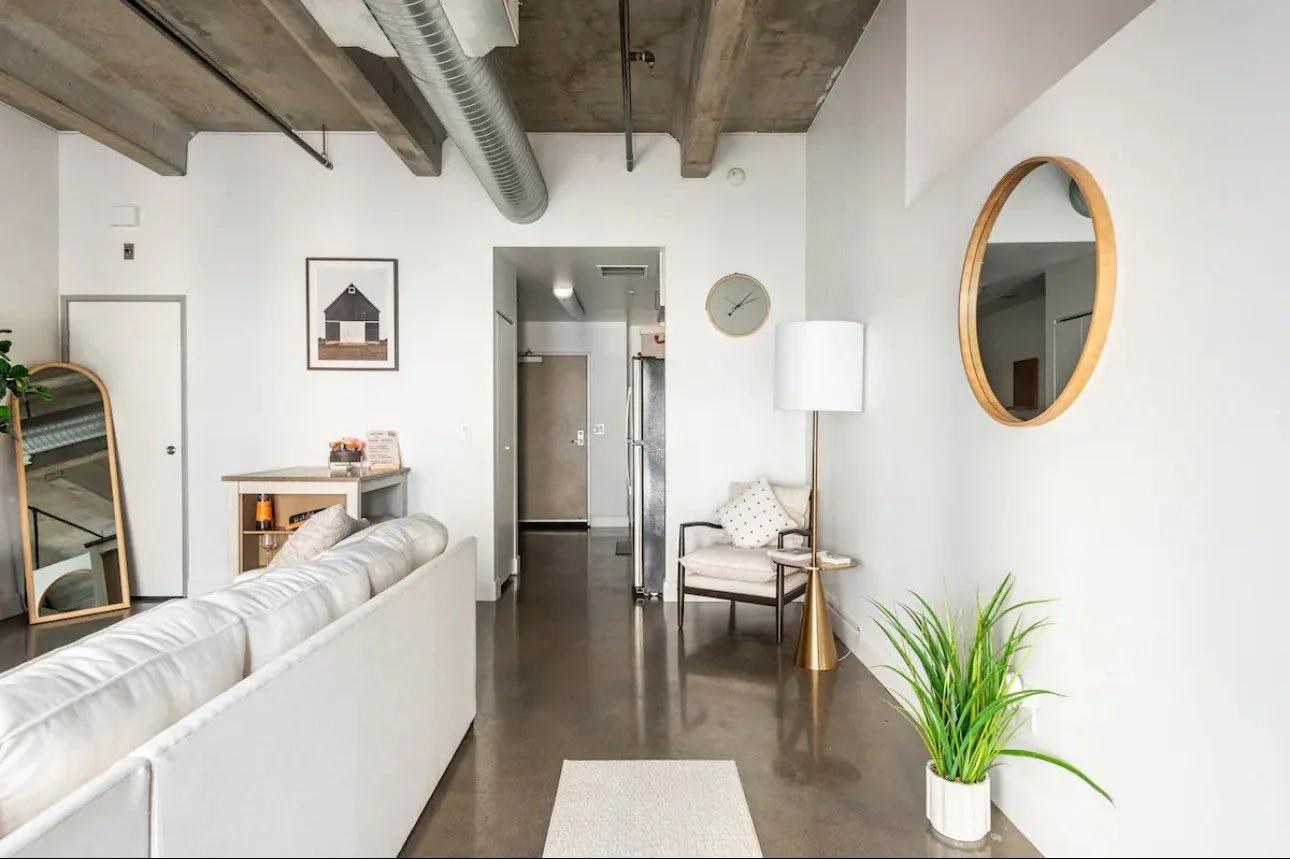 Modern living room with white walls, concrete ceiling, and polished concrete floor. Features a white sofa, a tall mirror, a potted plant, a floor lamp, and wall art. A doorway leads to a hallway with a clock on the wall.