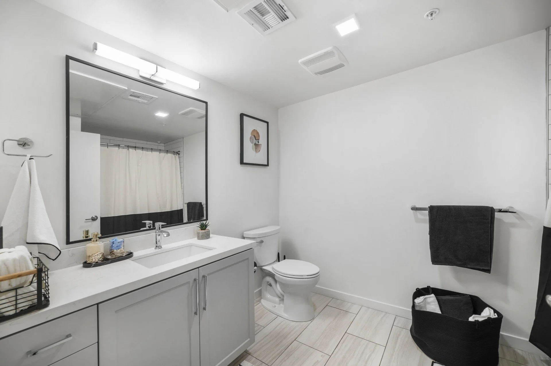 Modern bathroom with white walls, large mirror, vanity with soap dispenser and plant, toilet, black towel on rack, and black basket with towels.