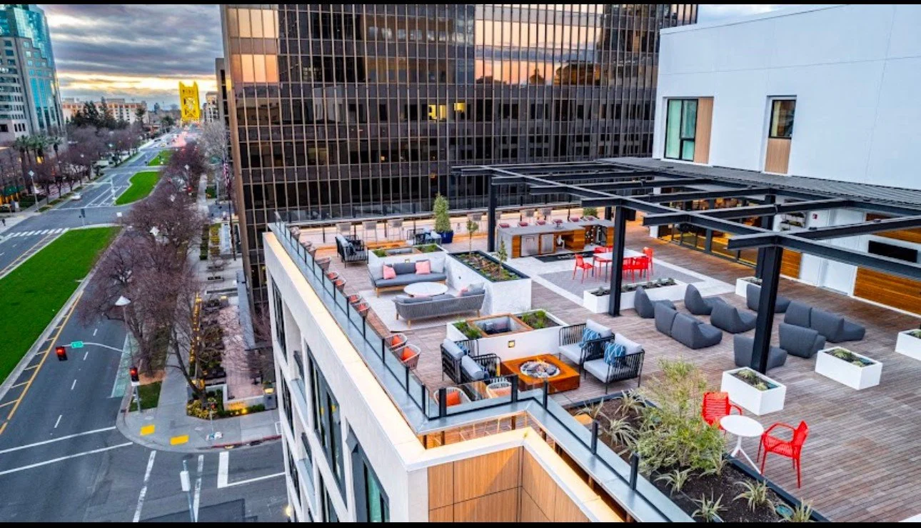 Rooftop terrace with seating areas, tables, and dining spaces, overlooking city streets and modern buildings at sunset.