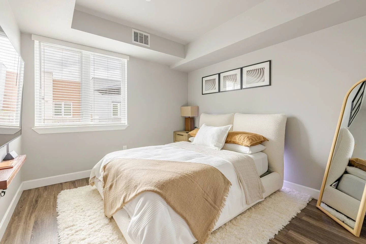 A bedroom with a white bed against a gray wall, framed artwork above, a beige nightstand with a lamp, large windows with blinds, hardwood floors with a cream rug, and a full-length mirror.