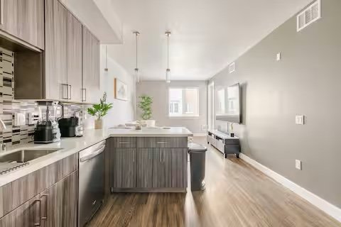 Open modern kitchen and living room with hardwood floors, gray cabinets, white countertops, and a TV on the wall.