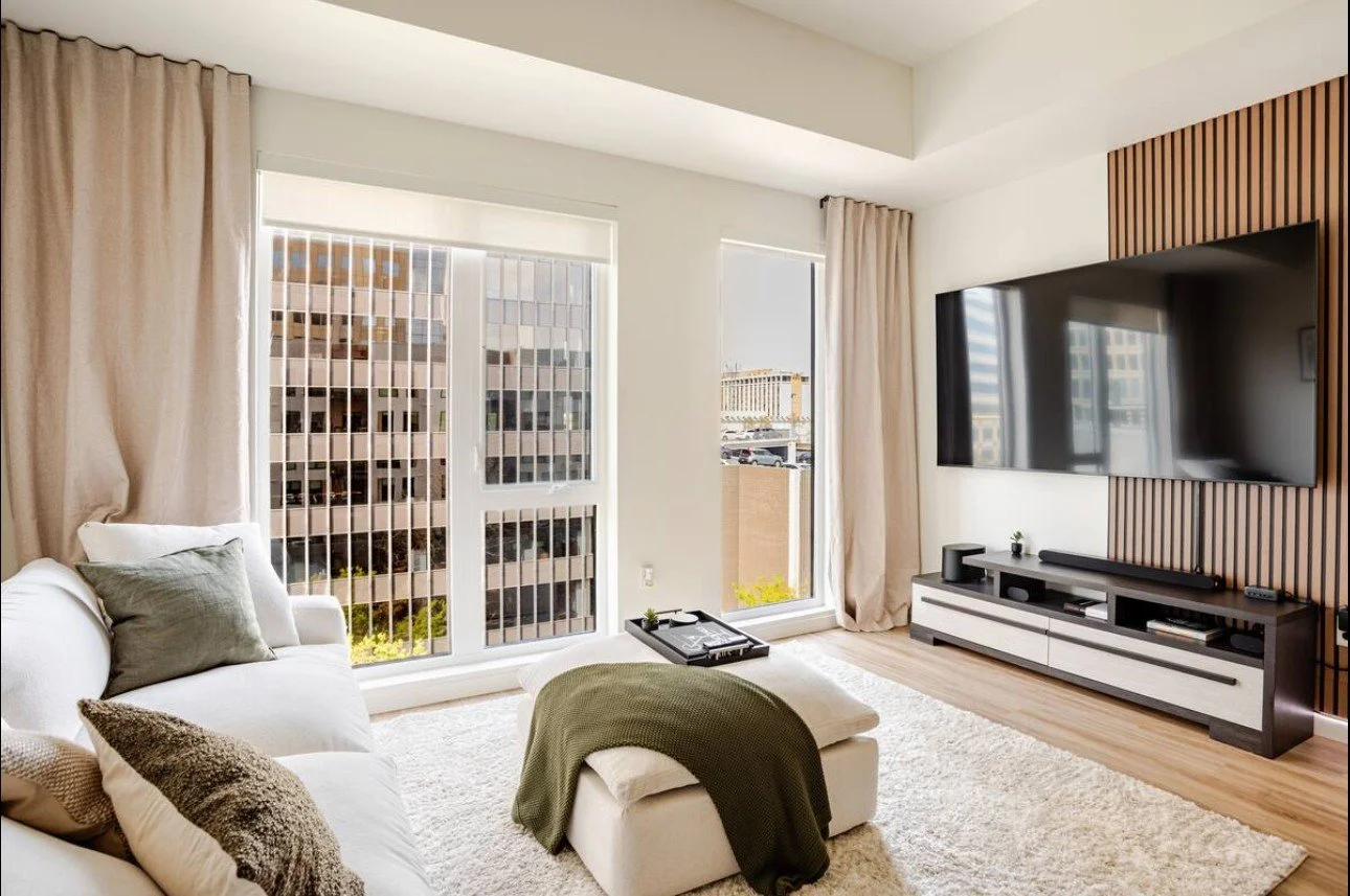 Modern living room with large windows, beige curtains, white sofa with gray and beige pillows, a beige ottoman with a green throw, a black and white entertainment center, a large flat-screen TV, and a wooden accent wall.