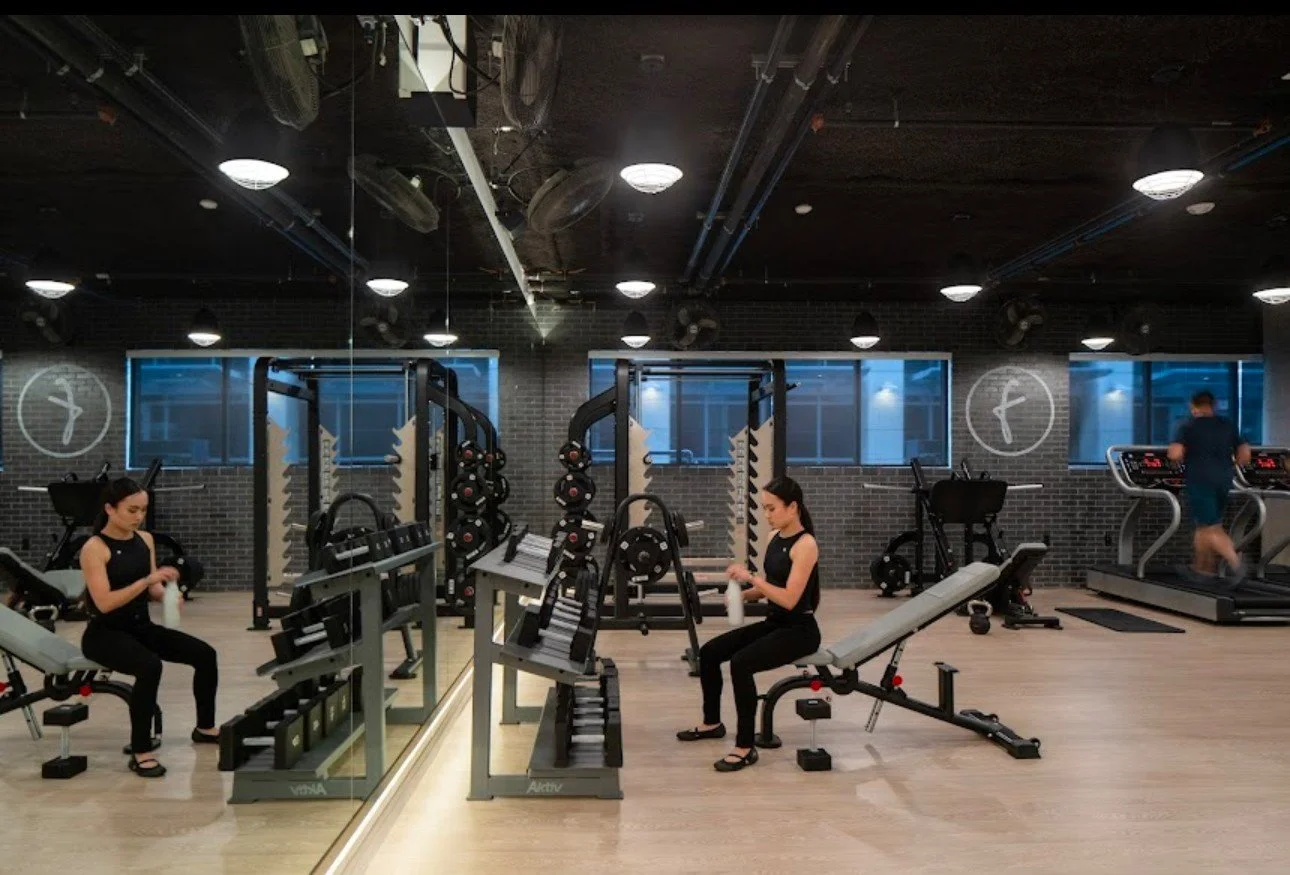 Women sitting on exercise benches, drinking water bottles, in a modern gym with cardio and weight equipment, and a man running on treadmill in the background.