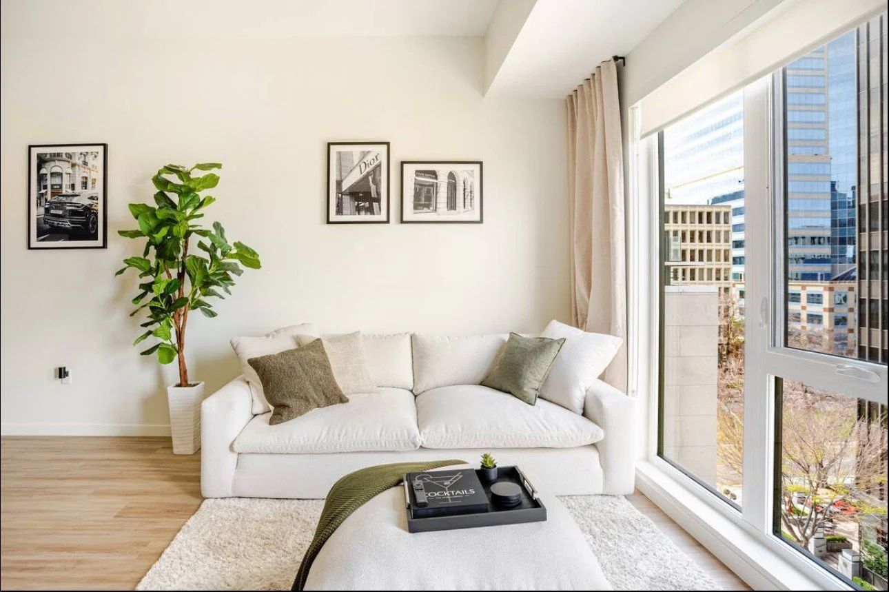 Modern living room with a white sofa, throw pillows, potted plant, framed wall art, large window with city view, beige curtains, and a black tray with drinks and decor on an ottoman.