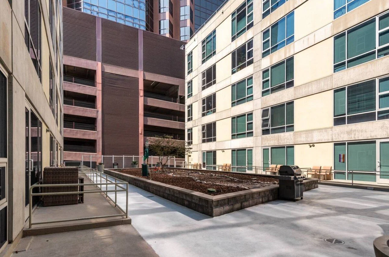 Empty outdoor courtyard between tall modern buildings with a small garden bed, benches, a barbecue grill, and a single tree.