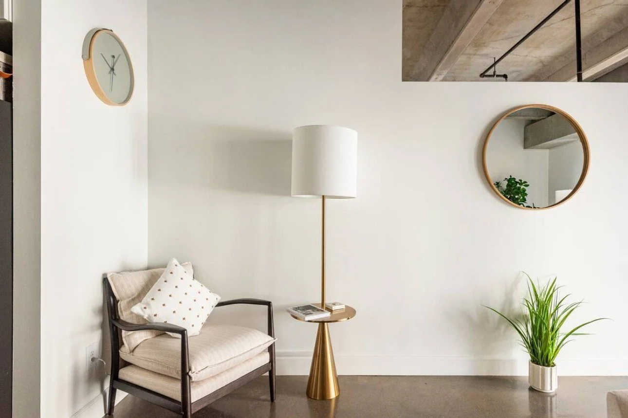 Minimalist living room corner with a beige armchair, white cushion, tall white floor lamp with gold stand, round mirror on white wall, and a potted green plant in a white pot.